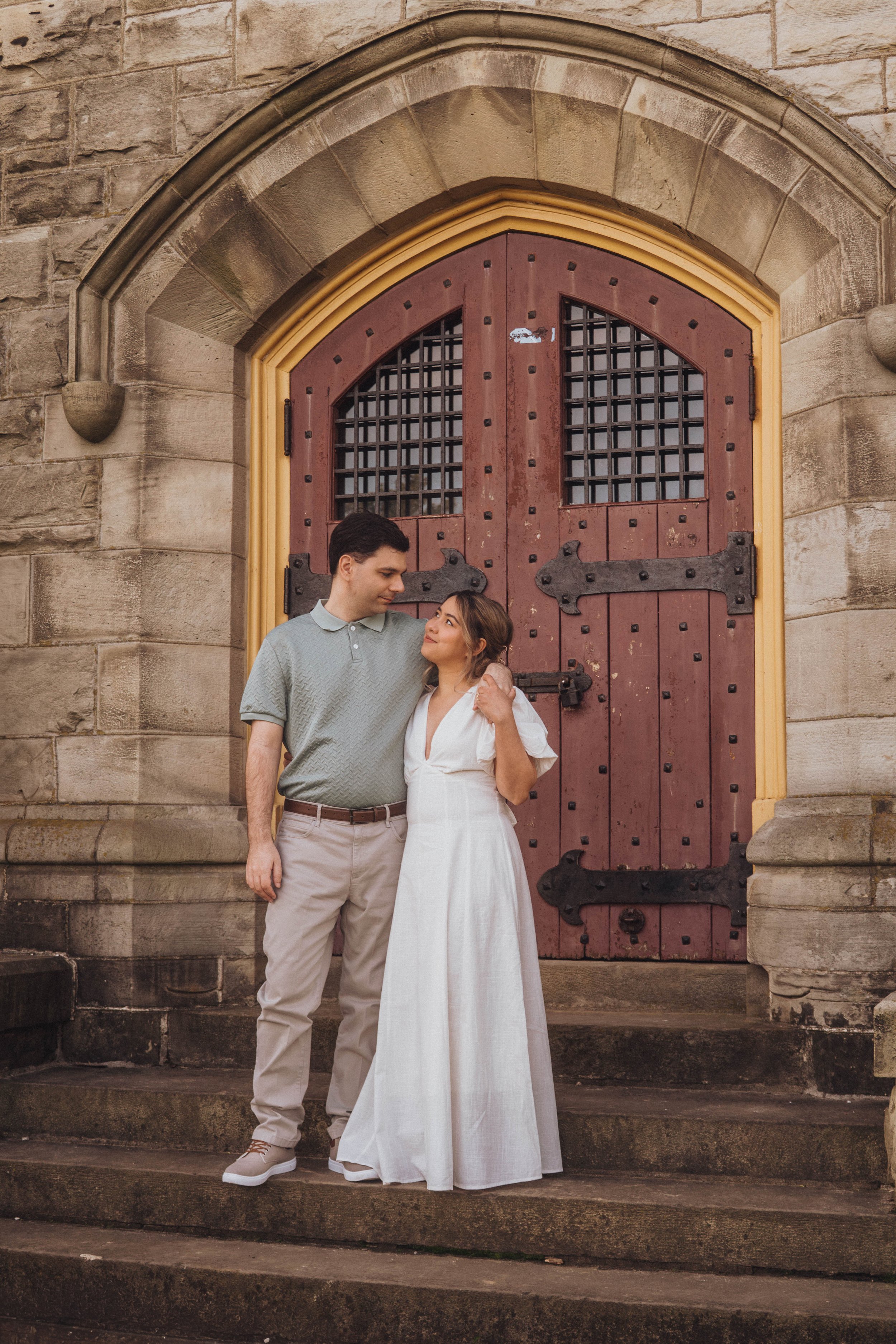 A young couple standing close together on the steps in front of a large, historic wooden door with iron hinges, surrounded by stone masonry. The man, wearing a light gray polo shirt and beige pants, gazes lovingly at the woman. The woman, in a flowin