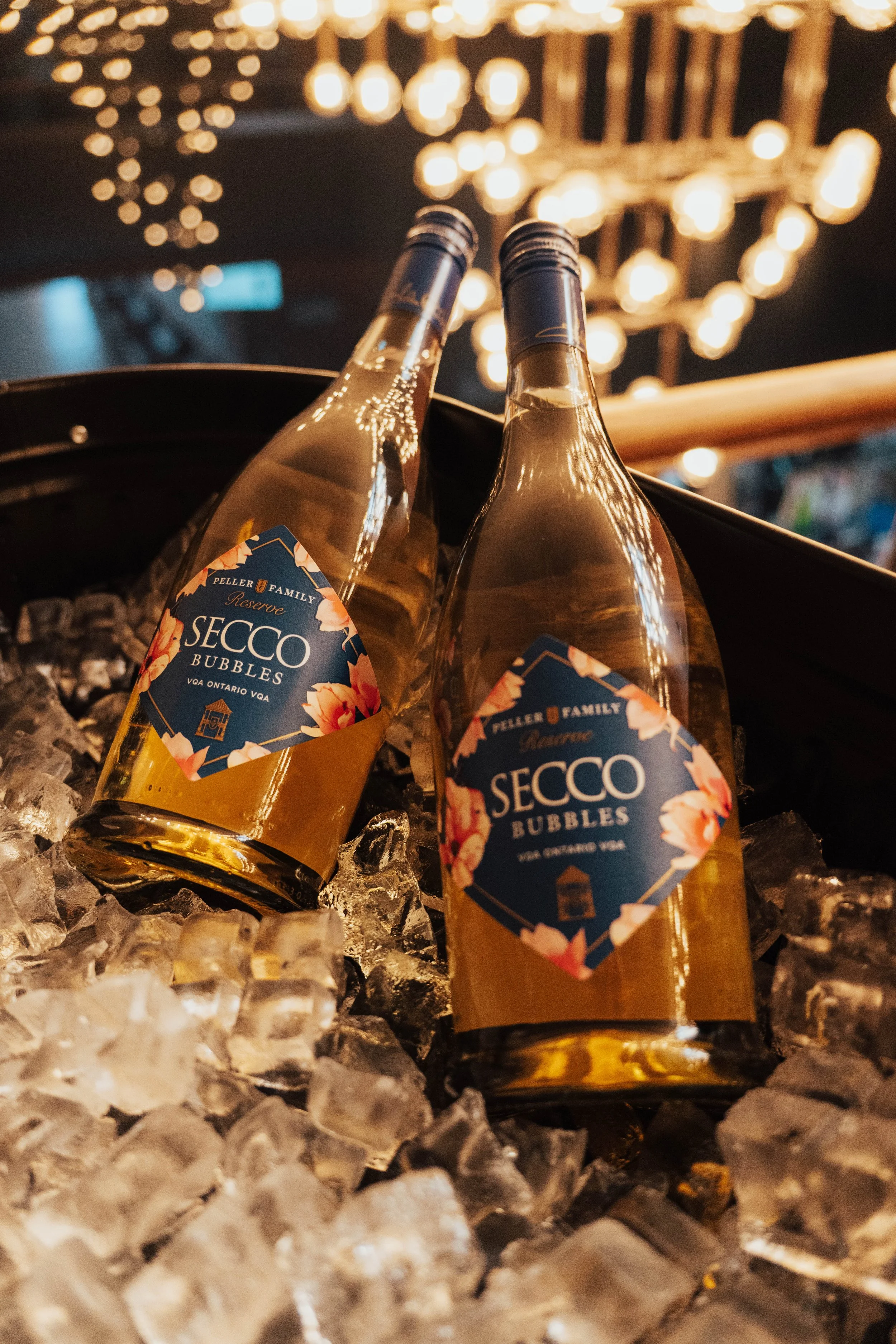 Two bottles of Secco Bubbles sparkling wine in an ice bucket filled with ice cubes, with a warm, indoor chandelier in the background.