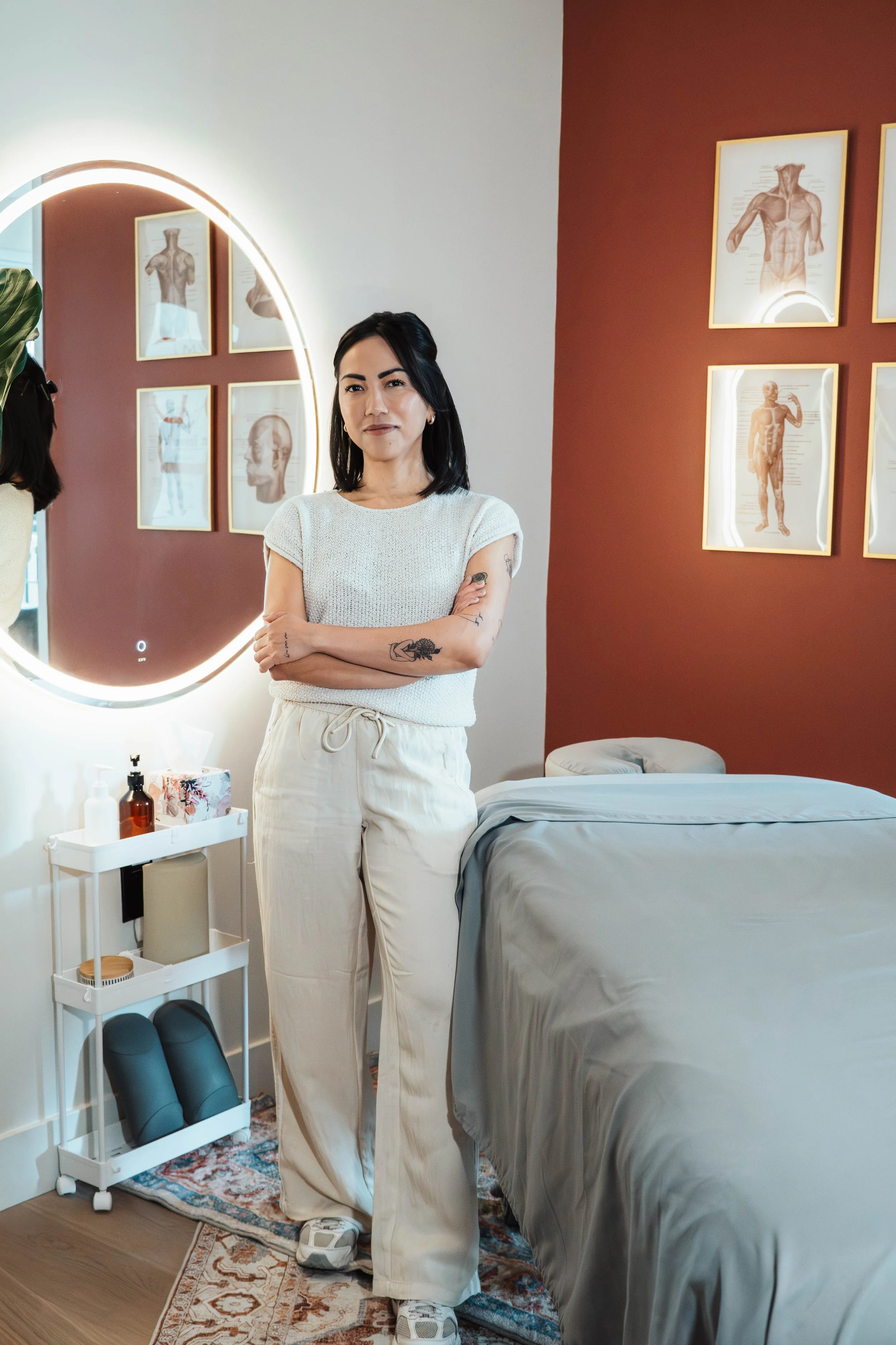 A woman with black hair and tattoos on her arms standing in a wellness or massage room. She is wearing a white top and beige pants, with her arms crossed. The room has framed anatomical posters on the wall, a glowing mirror, a small cart with bottles