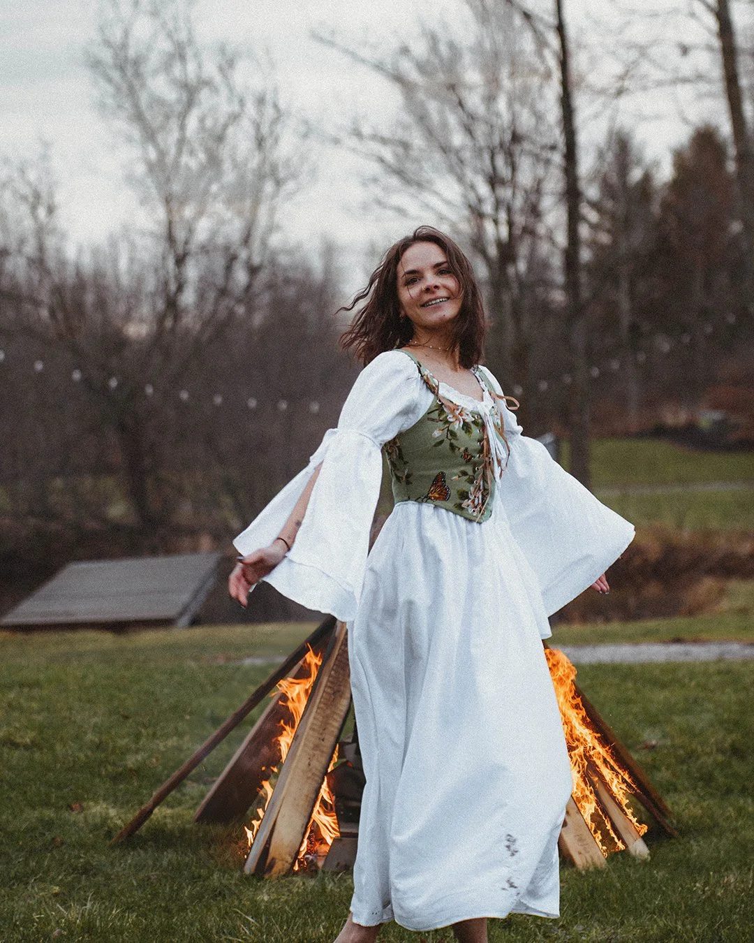 A woman in a white dress and a green embroidered vest standing outdoors near a bonfire, with trees and a cloudy sky in the background.