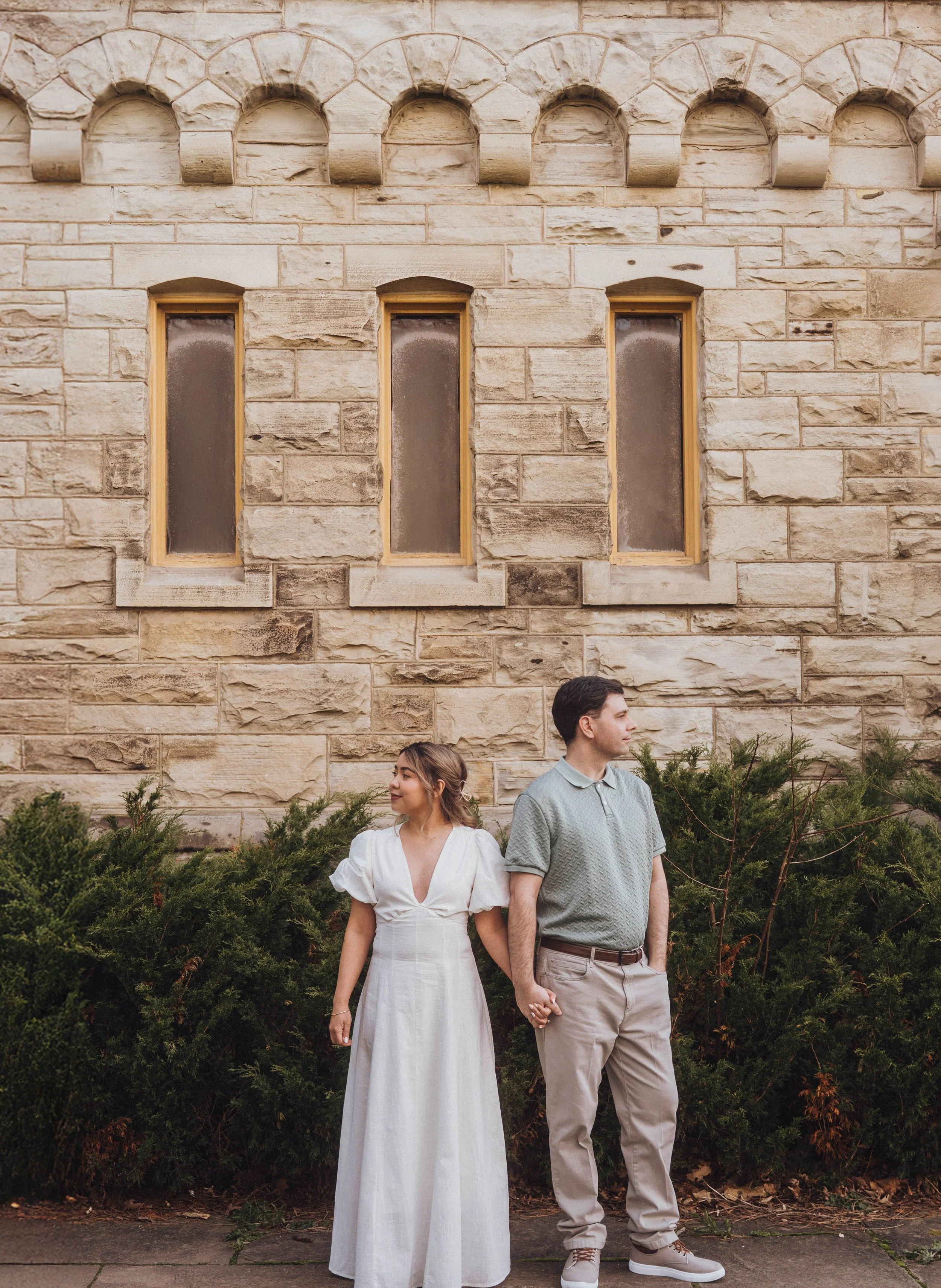 A young couple holding hands and standing in front of a stone wall with arched windows. The woman is wearing a white dress with puffed sleeves and the man is wearing a gray polo shirt and beige pants.