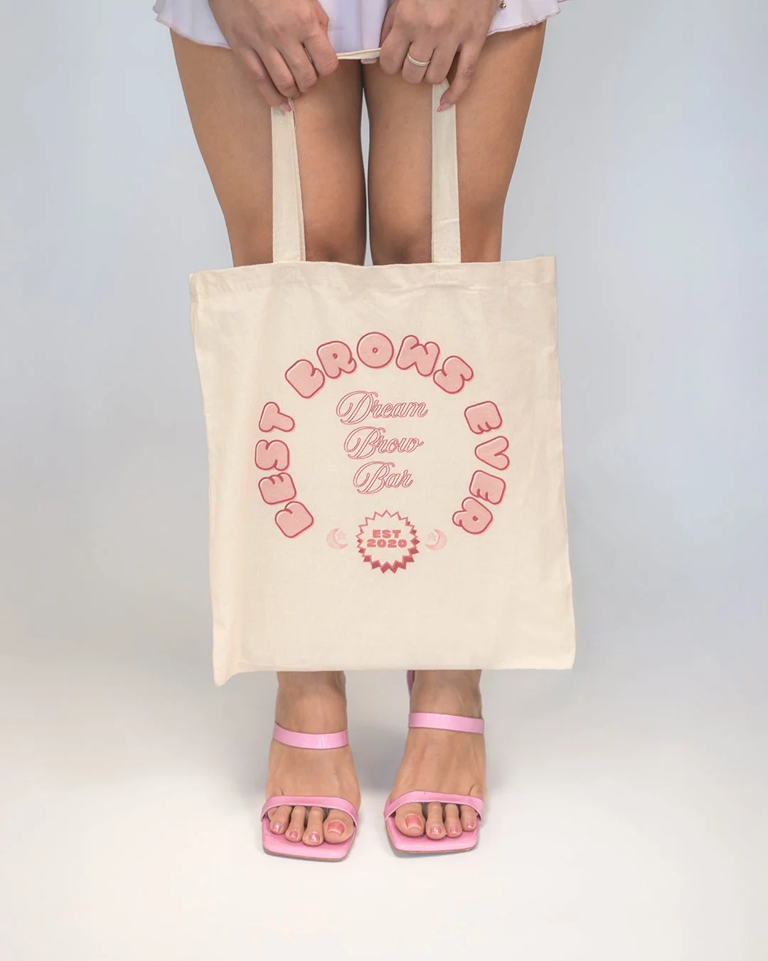 A person is holding a beige tote bag that reads 'Dream Brew Bar' and 'EST 2020' with pink decorative text and small heart design. The person is wearing pink high-heeled sandals and a short white dress, and their hands are visible at the top of the im