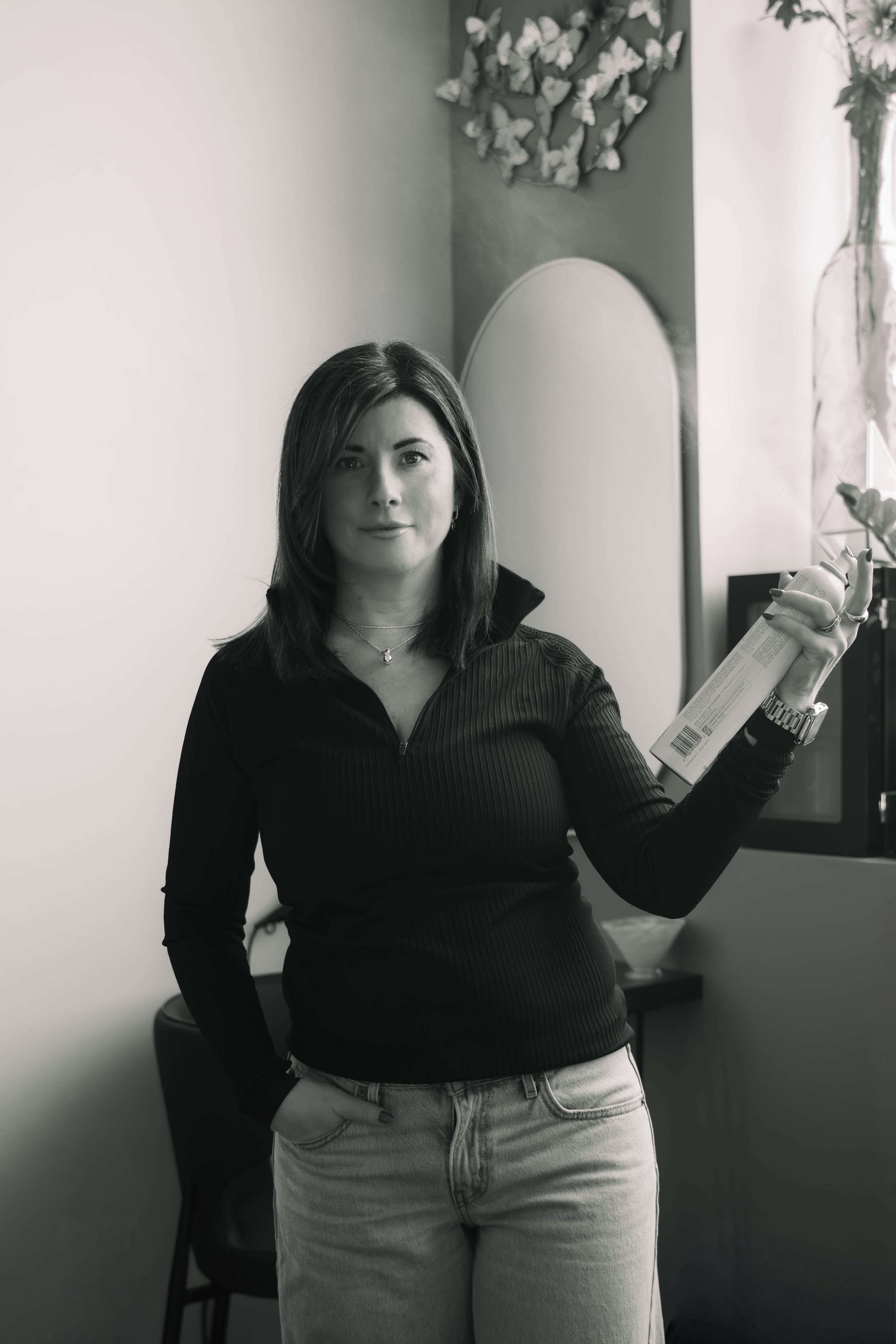A woman standing indoors holding a can of spray in her right hand. She is wearing a black zip-up top and light-colored jeans, with her left hand in her pocket. The background features a mirror, a decorative piece with butterflies on the wall, and som
