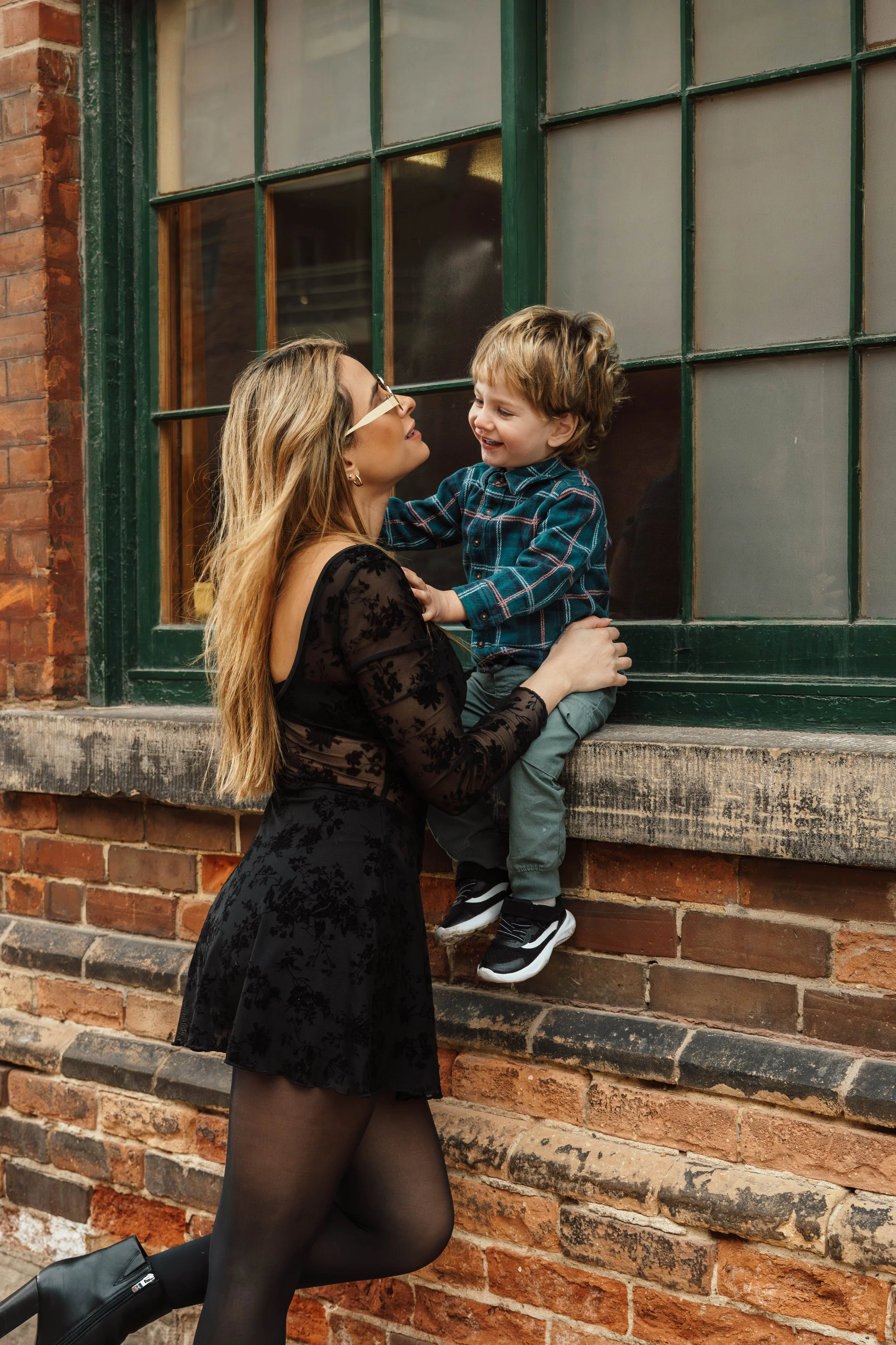 A woman dressed in black with sheer floral lace sleeves is kneeling on one knee on the sidewalk, holding a smiling young boy in a blue plaid shirt and grey pants near a large window of a brick building.