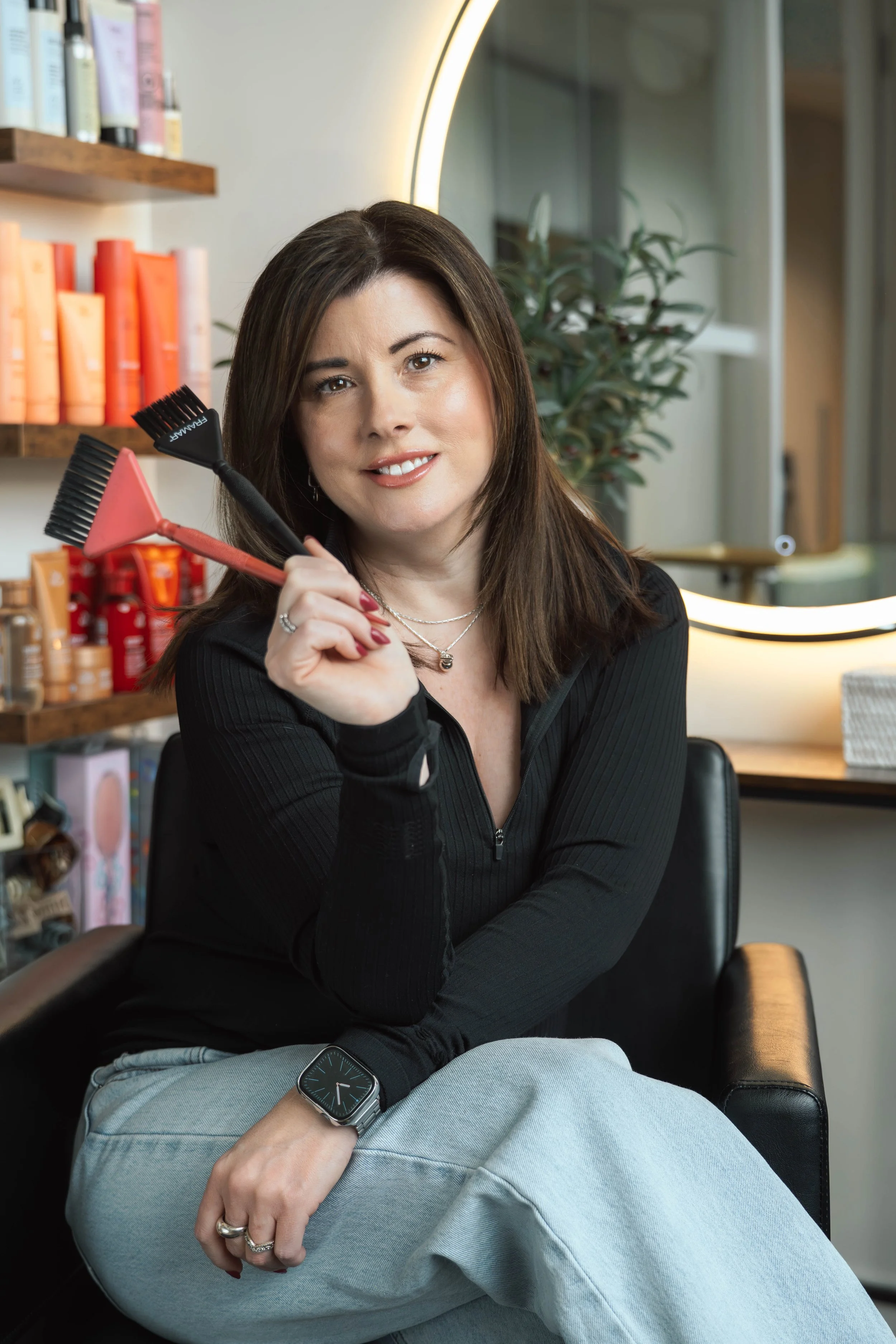 Woman sitting in a salon holding hair coloring brushes, with a mirror and hair products in the background.