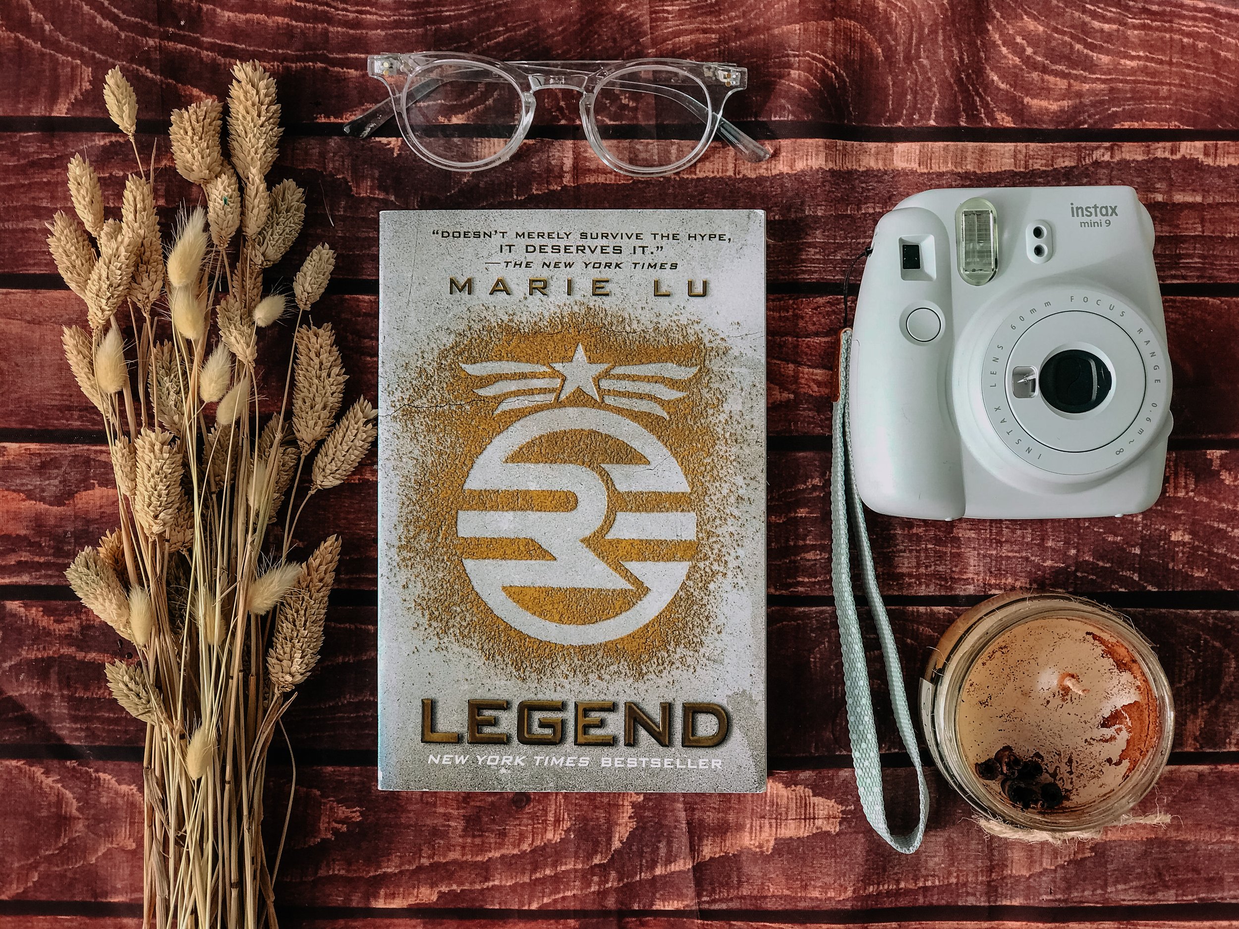 Flat lay of a dried wheat bouquet, eyeglasses, a book titled 'Legend' by Marie Lu, a white Instax mini 9 camera, and a glass of iced coffee with dark berries, all on a wooden surface.