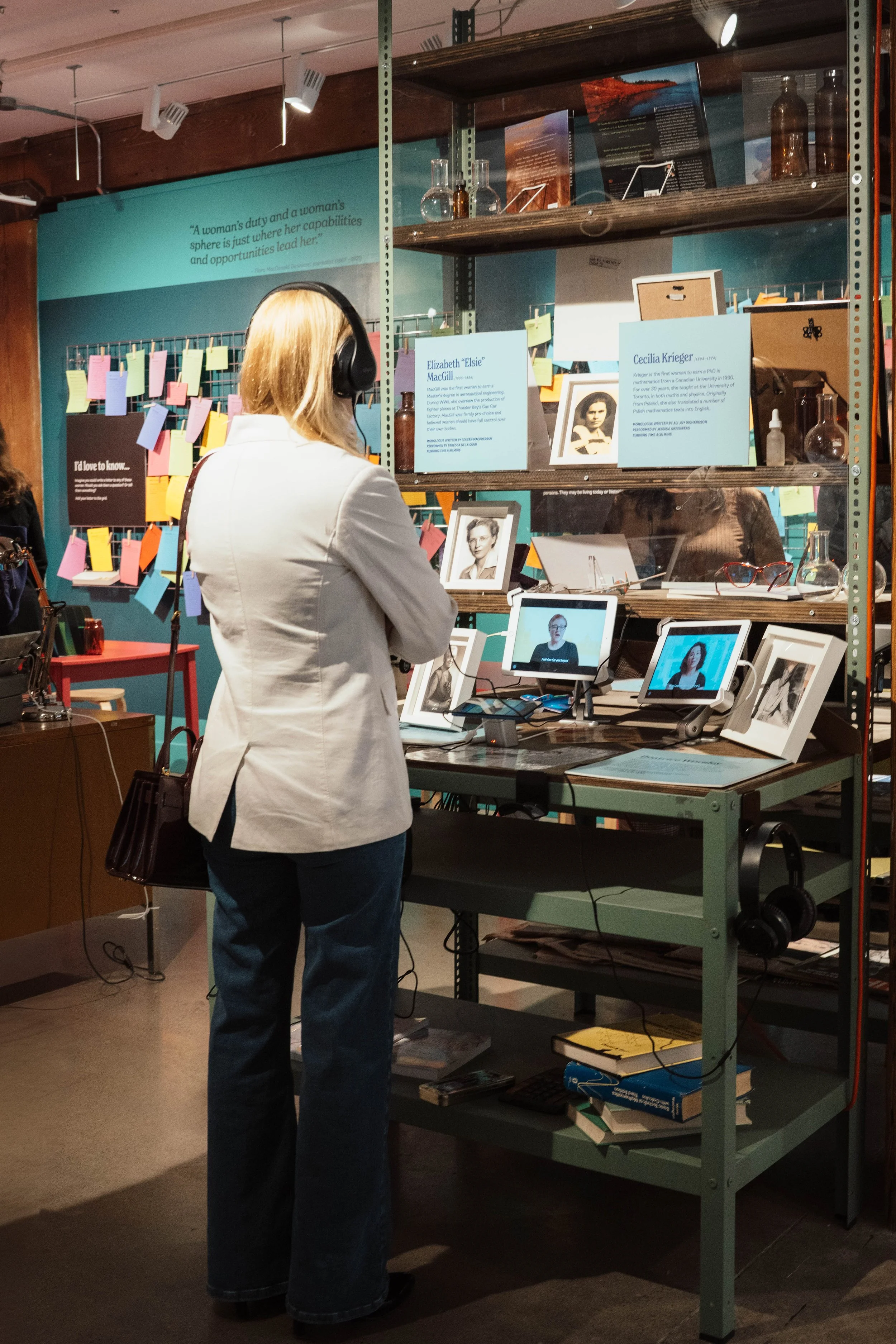 A woman with blonde hair wearing a white blazer and headphones stands in front of a display table with framed photographs and digital screens, in a museum or exhibit setting.