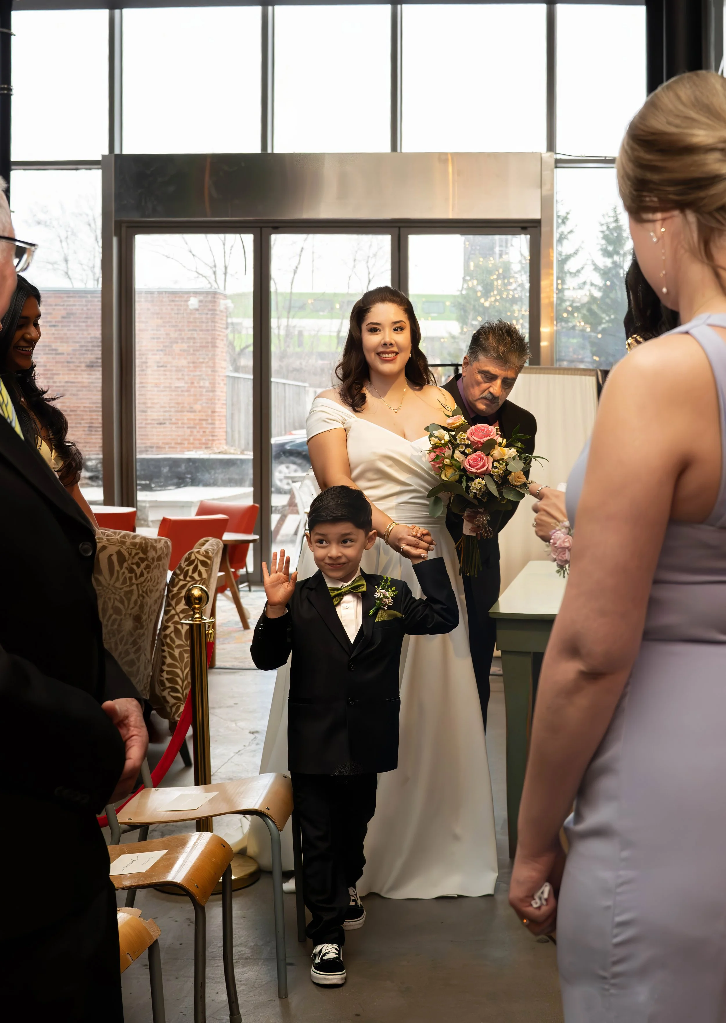 A bride in a white dress holding a bouquet of pink and white roses, smiling as she stands among wedding guests inside a venue with large glass windows showing an outdoor scene. A young boy in a black tuxedo with a green bow tie is in front, waving an