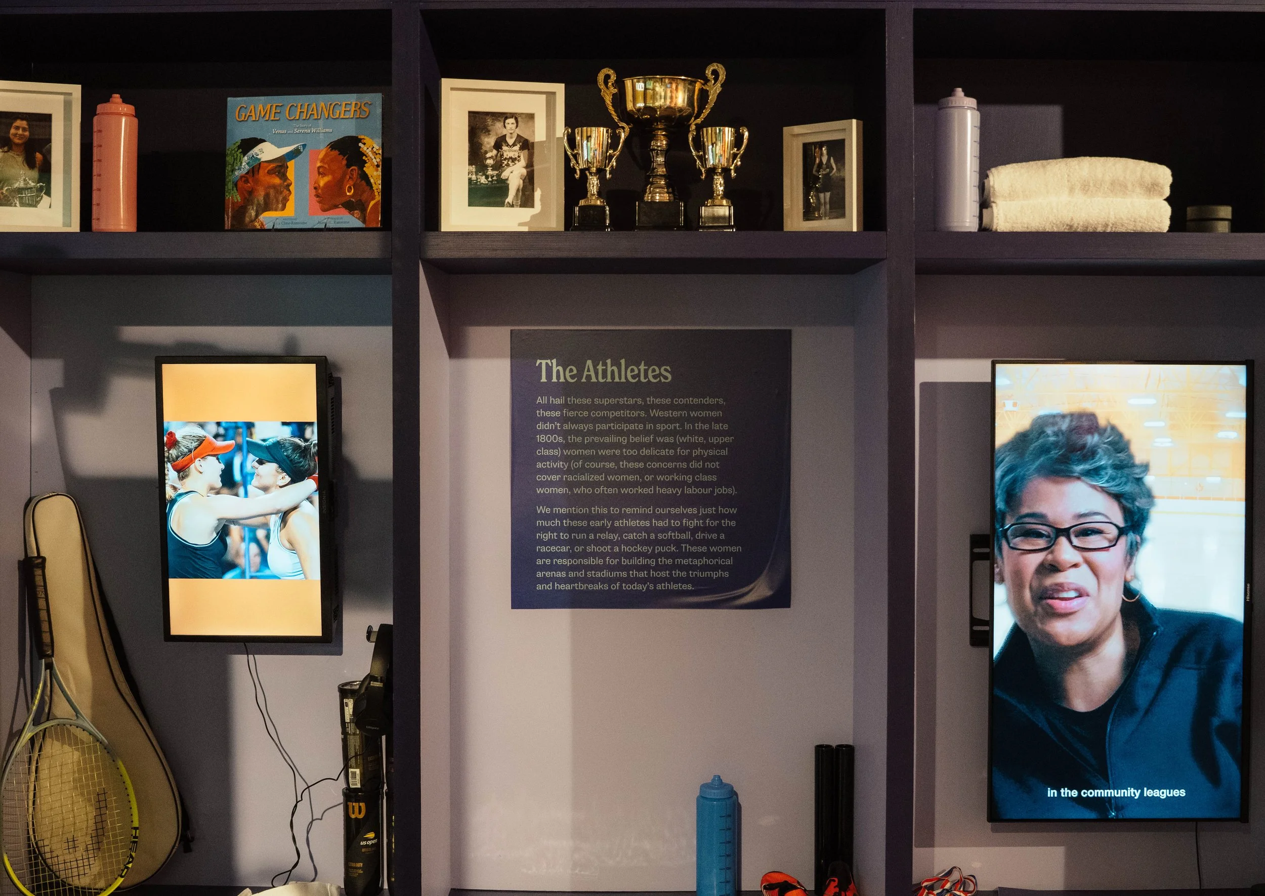 Display shelf with trophies, framed photos, water bottles, rolled towels, and digital screens featuring people playing tennis and a woman speaking, with a text panel titled 'The Athletes' about female athletes' history and challenges.