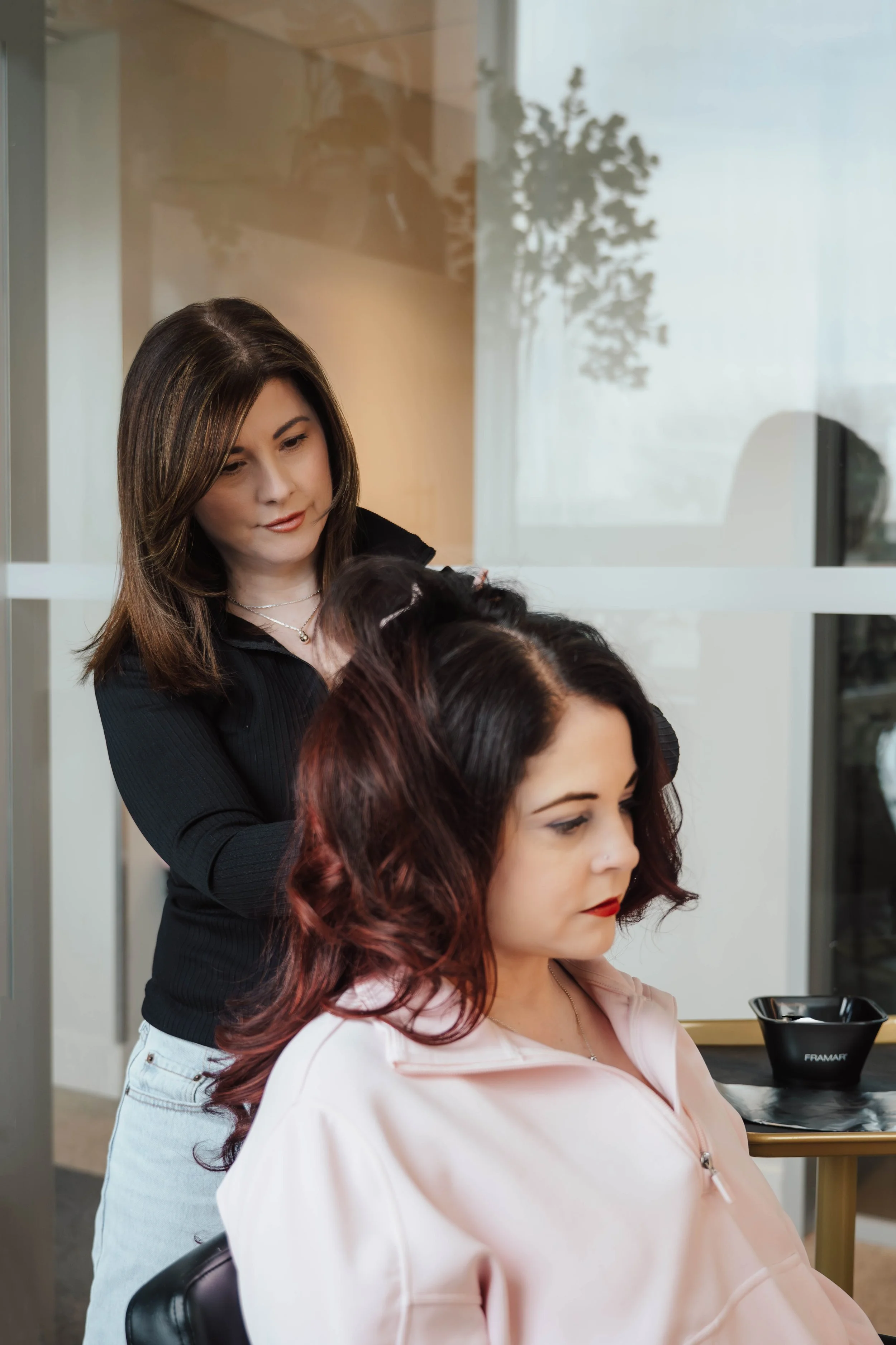 A hairstylist is styling a woman's hair in a salon. The woman is seated wearing a light pink top, and the hairstylist is standing behind her, working on her hair.