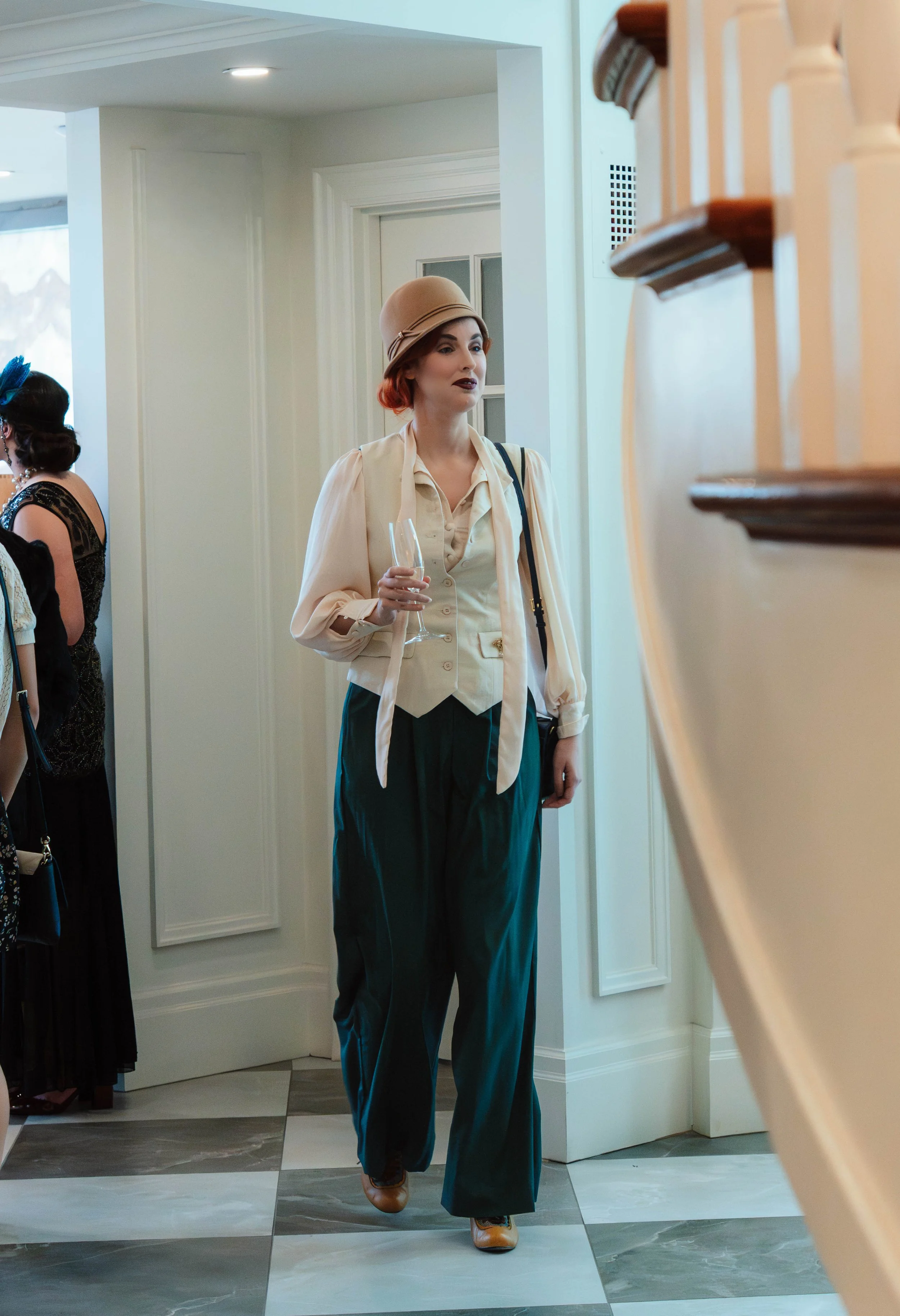A woman dressed in vintage style clothing, wearing a beige hat, off-white blouse, vest, and dark green wide-leg pants, holding a champagne flute, standing inside a house with white walls and a staircase.