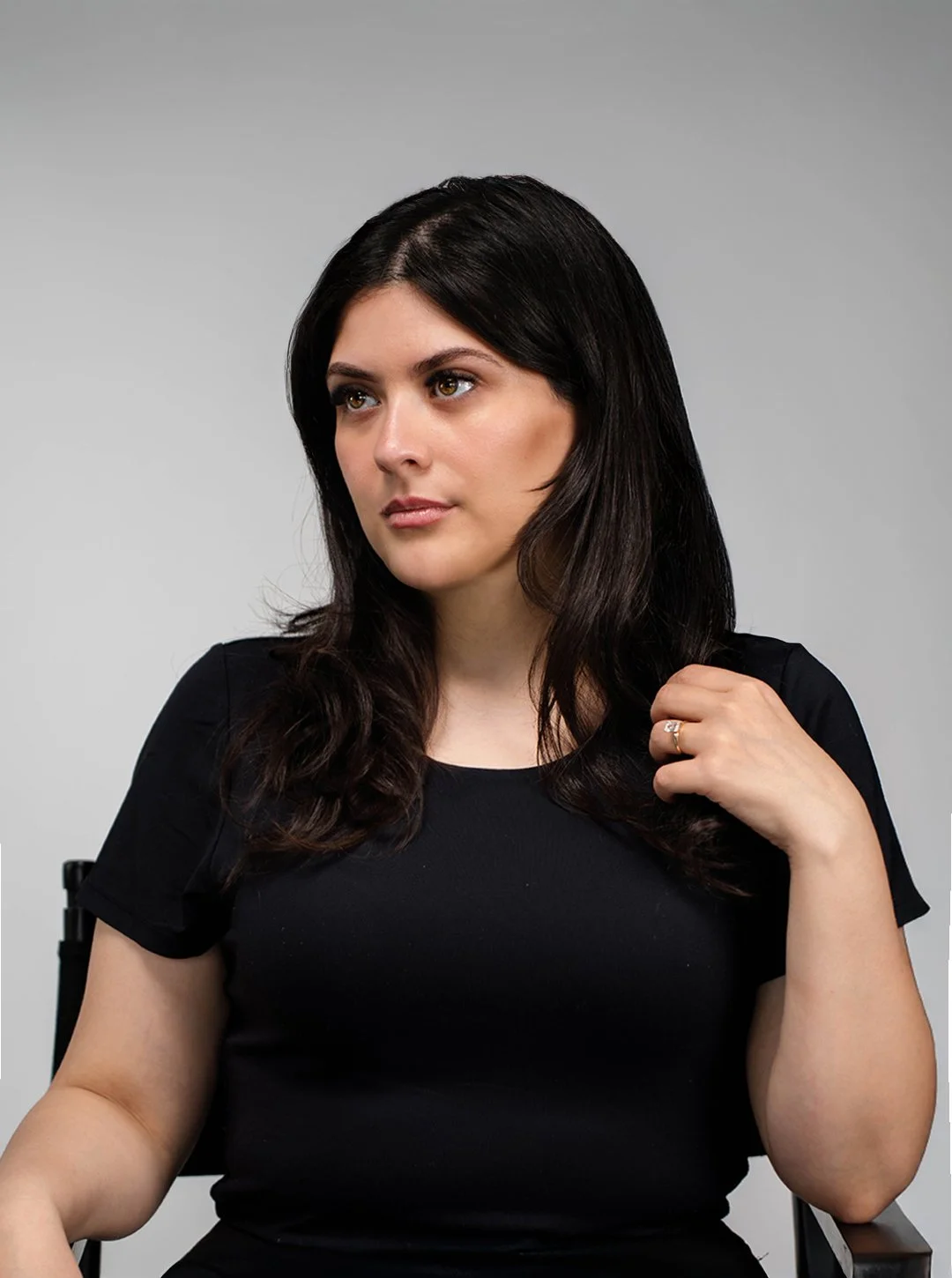 A young woman with dark hair and light skin tone, wearing a black T-shirt, sitting in a chair against a plain gray background. She is looking to the side with a neutral expression and touching her hair with her right hand.