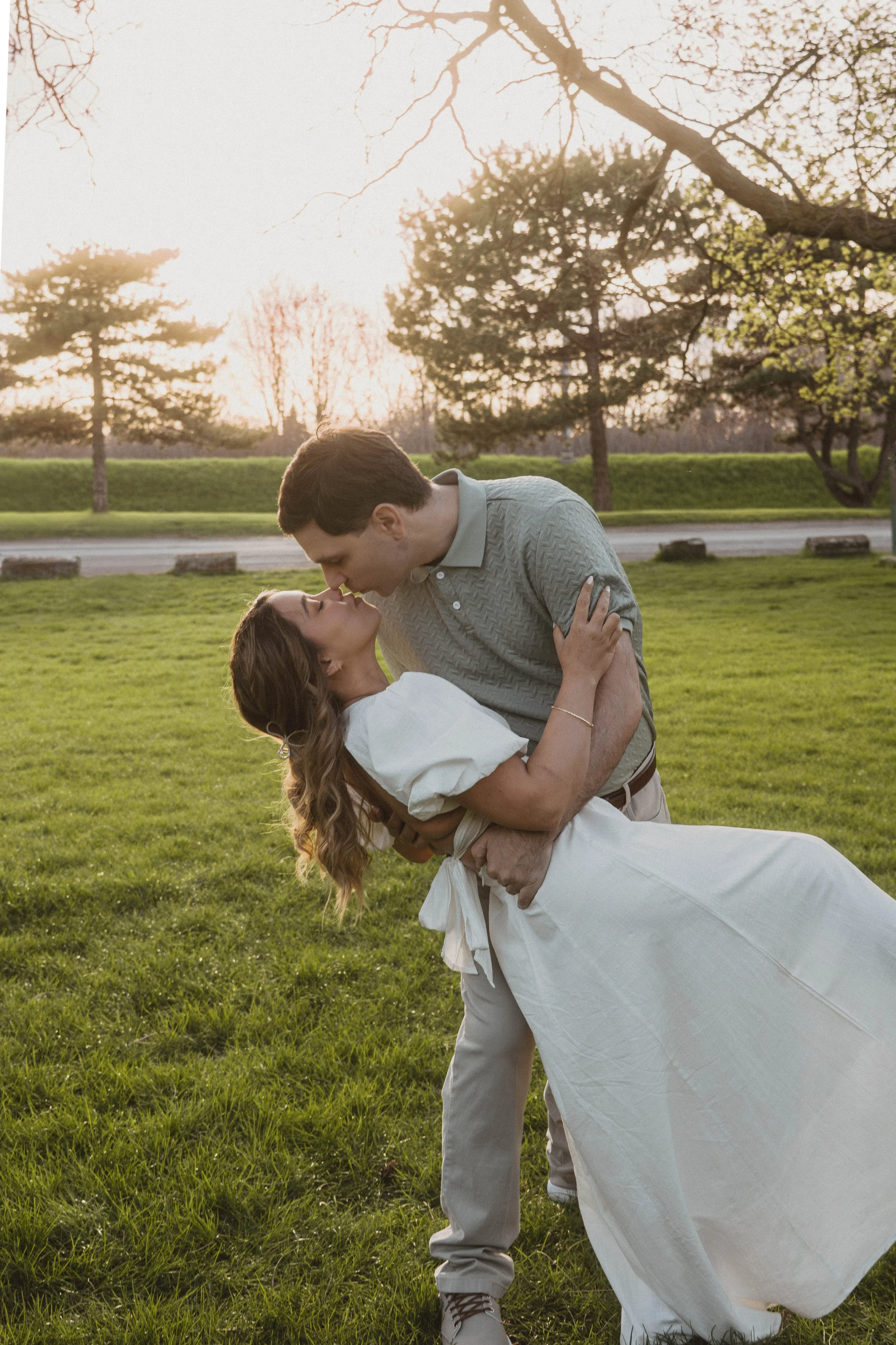 A couple in a park during sunset, with the man lifting and kissing the woman who is in a white dress, holding onto him with an expression of happiness.