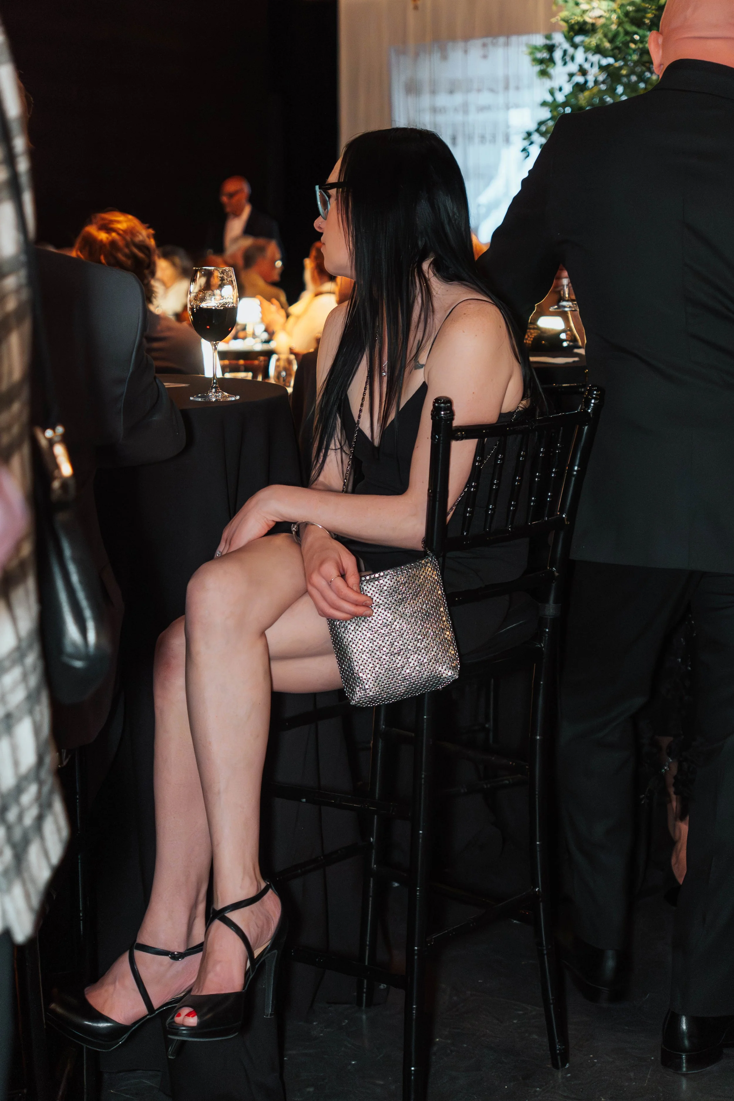 A woman with long black hair, glasses, and a black dress sitting at a table with a glass of red wine, surrounded by other people at a formal event.