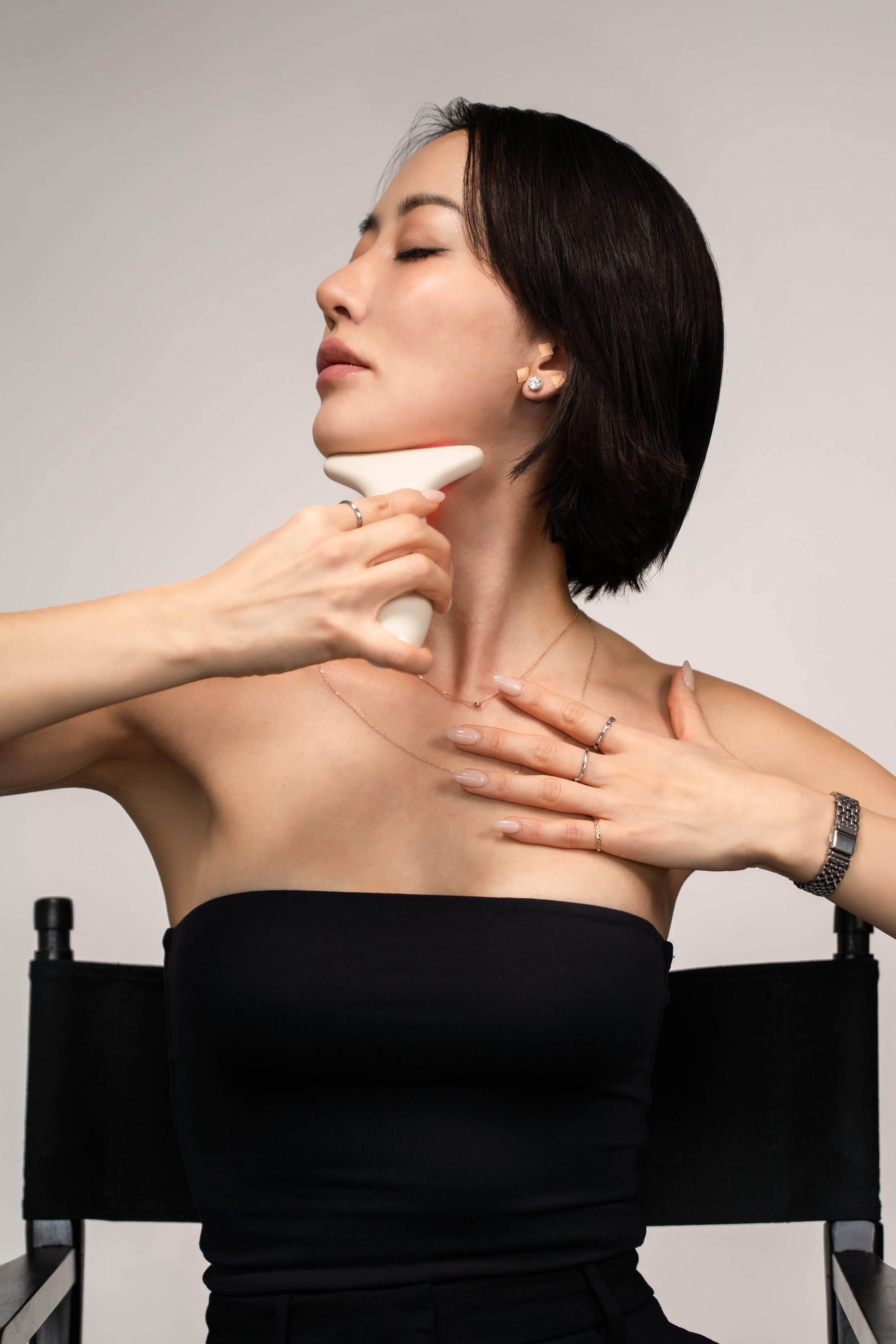 A woman sitting on a chair having her neck examined with a skincare device, wearing a strapless black top, jewelry, and a watch.