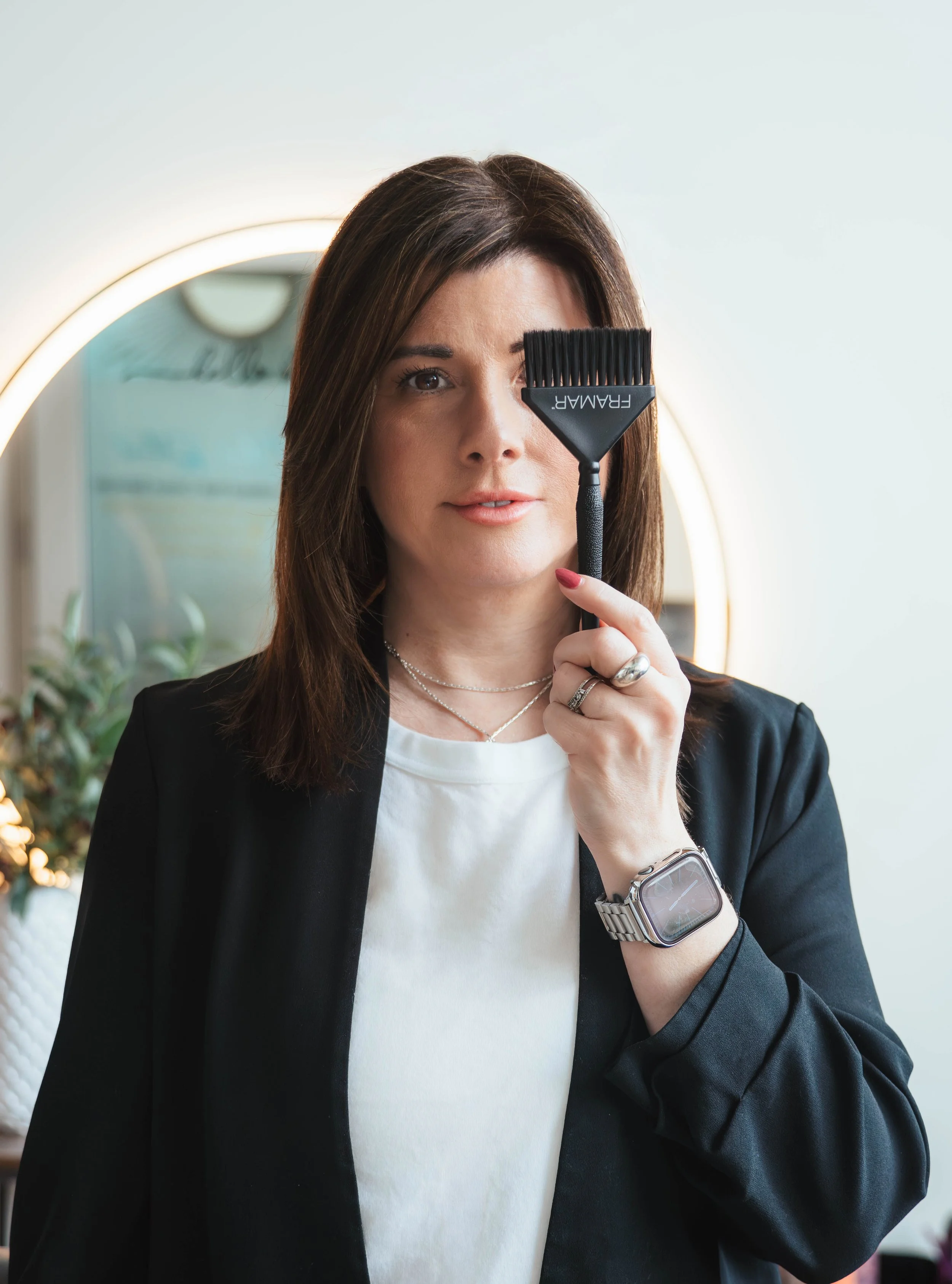 A woman with shoulder-length brown hair holding a black scalp brush near her eye, standing in front of a round mirror with light around it, wearing a black blazer, white shirt, a few necklaces, a silver watch, and rings on her fingers.