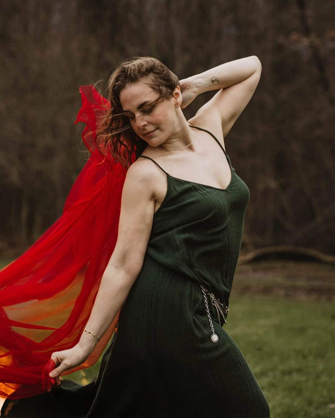 A woman with shoulder-length brown hair, wearing a dark green dress with thin straps, is outdoors with a blurred natural background. She is holding a red sheer fabric behind her, and her eyes are closed with a gentle expression.