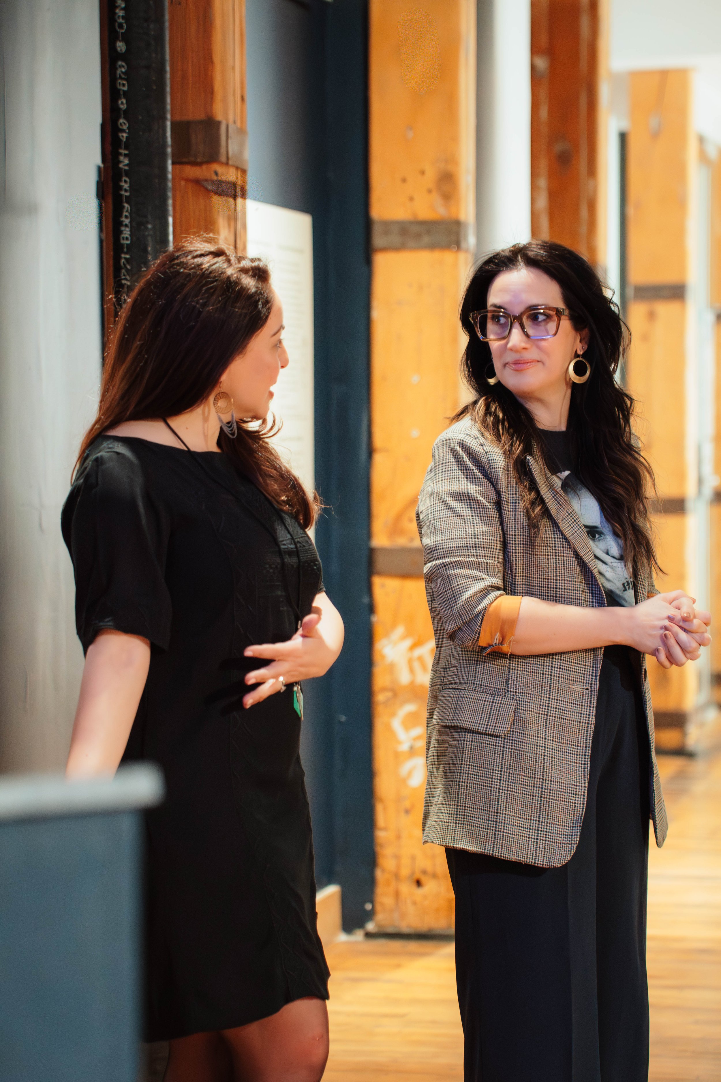 Two women standing and conversing indoors, one wearing a black dress and the other wearing a plaid blazer and glasses, with wooden beams and graffitied walls in the background.