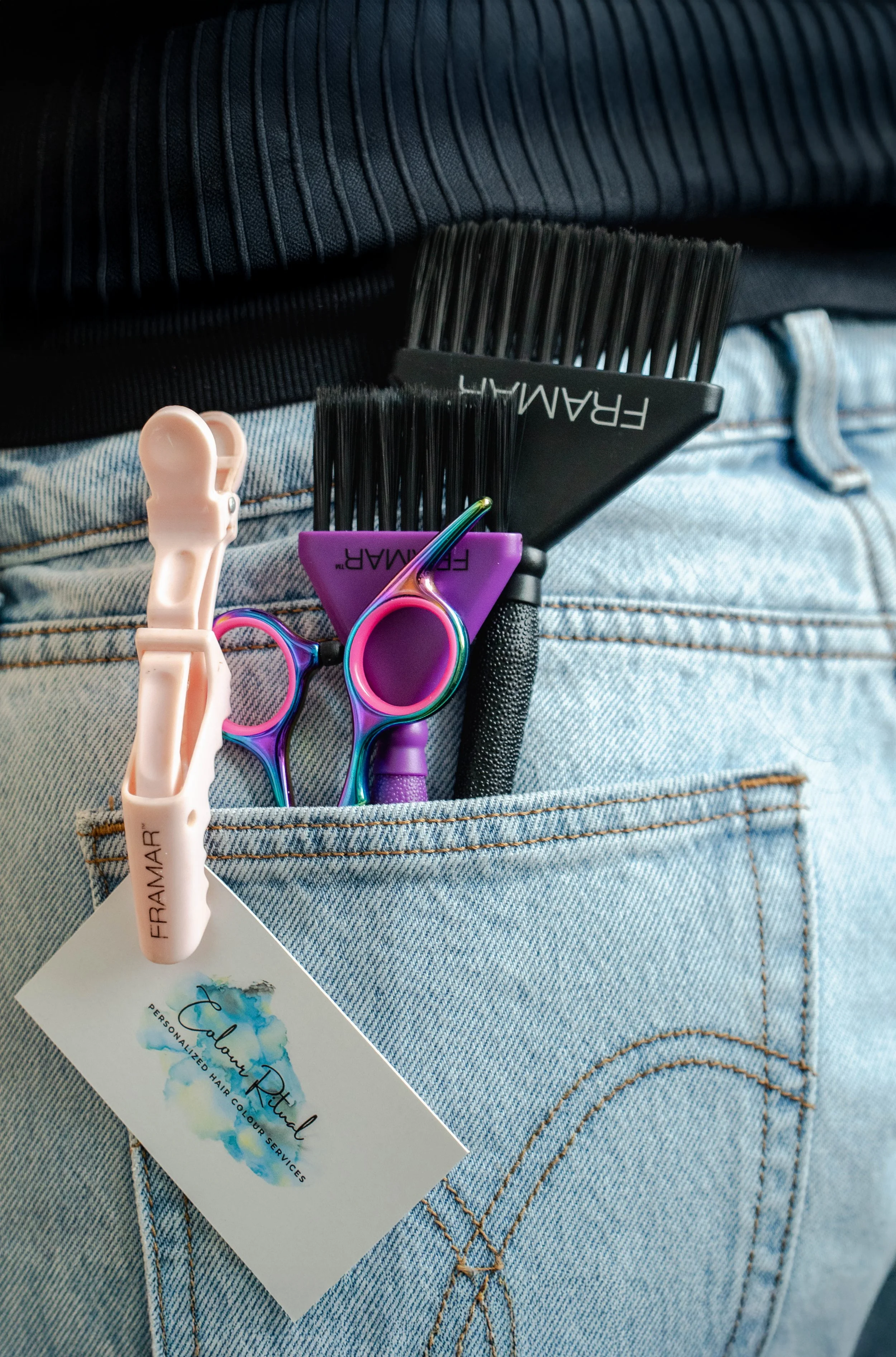 A close-up of a person's back pocket with hair cutting tools including scissors, small brushes, and a clipper, tucked into the pocket of light blue jeans.