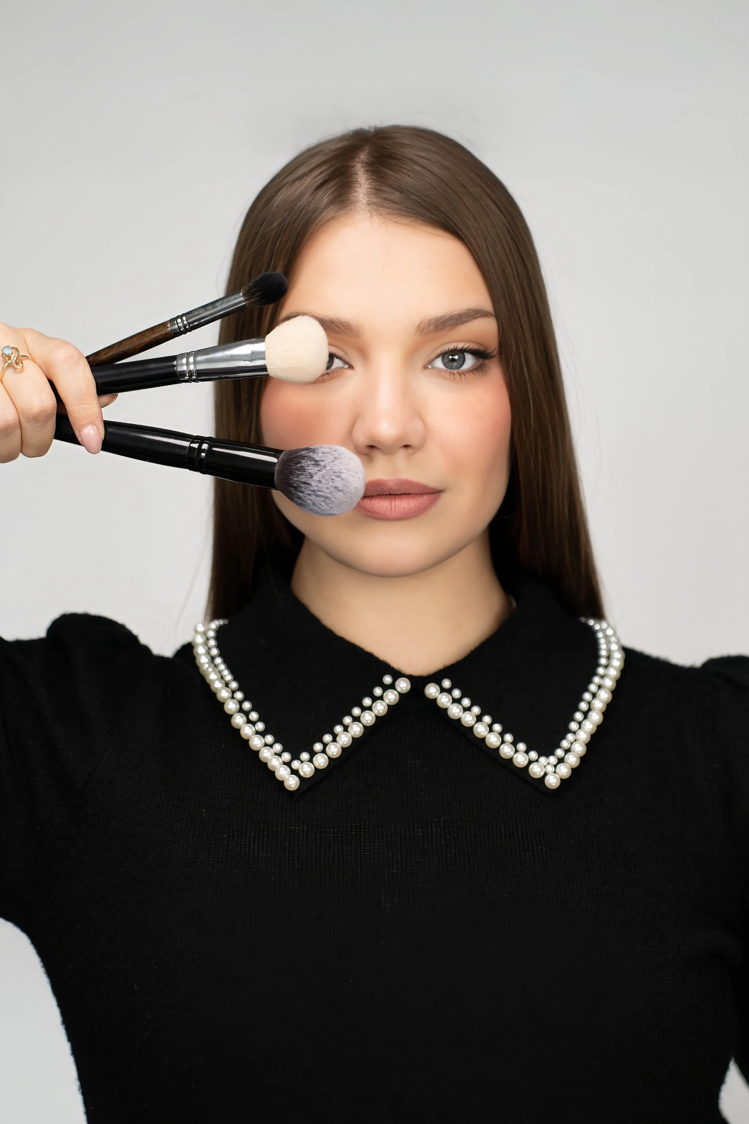 A woman with brown hair and fair skin holding three makeup brushes near her face.
