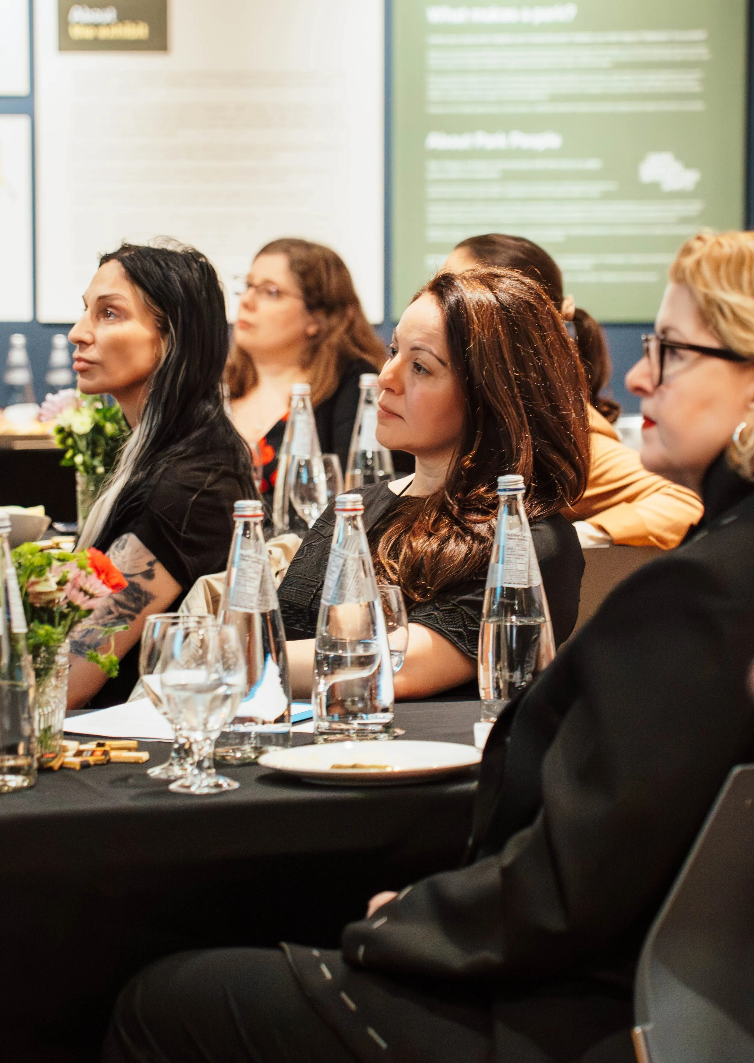 Women sitting attentively at a conference or seminar, with glass bottles of water, glasses, and flowers on the table in front of them.