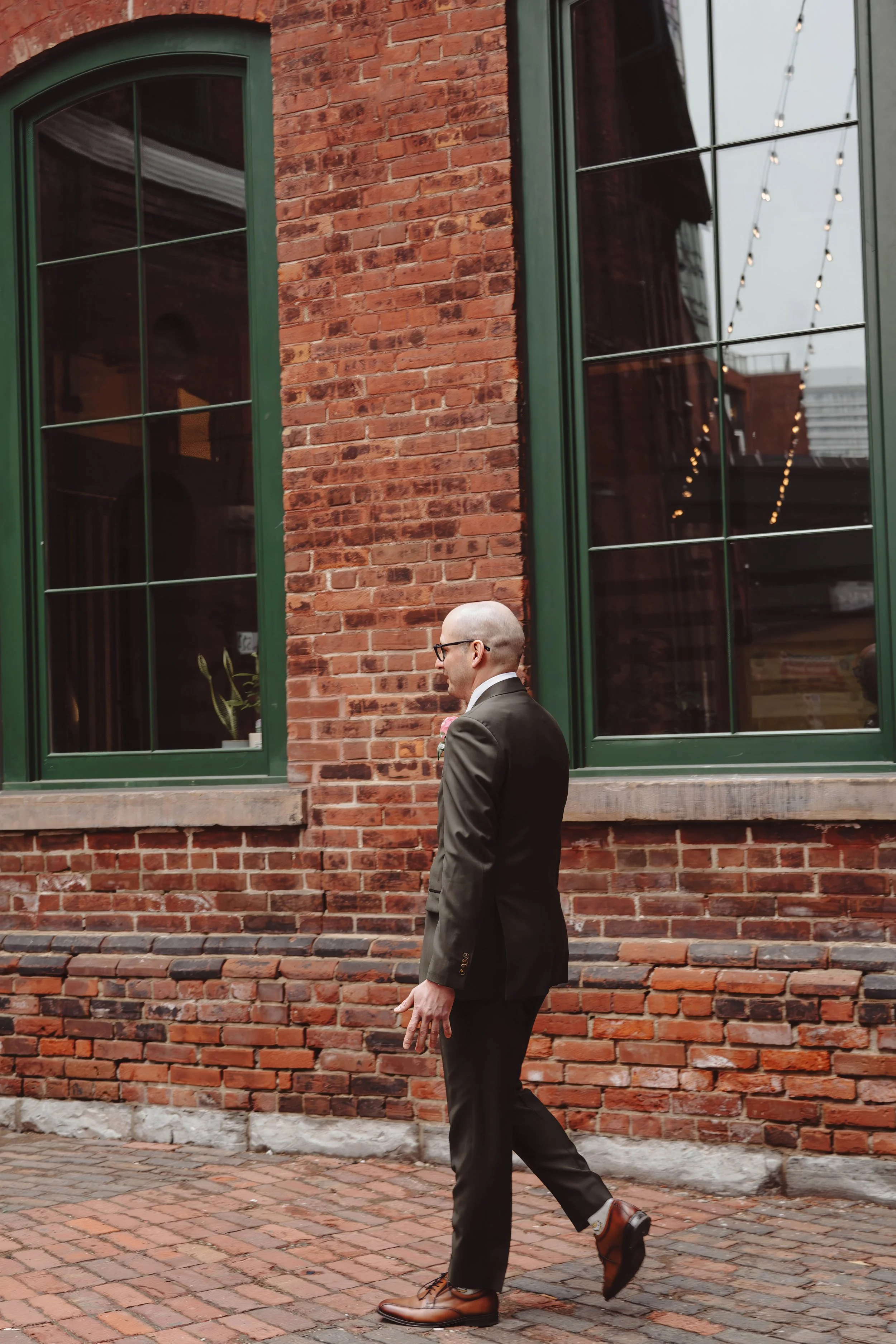 A man in a black suit walking past a brick building with green window frames.