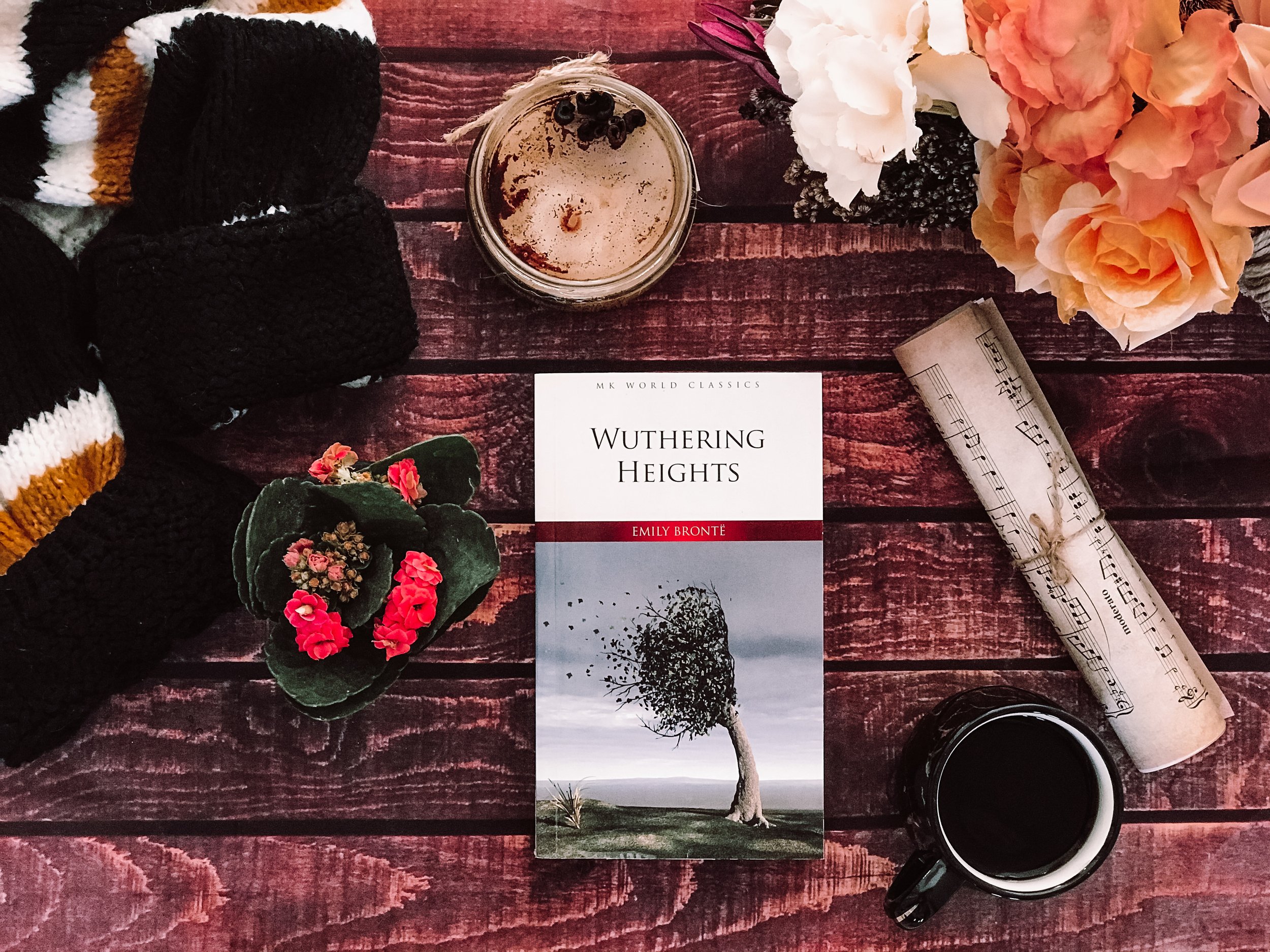 A flat lay of a red wooden table with a copy of Emily Brontë's novel "Wuthering Heights" in the center, surrounded by a cup of coffee, a glass jar with a beverage topped with black berries, a bundle of sheet music tied with string, a black mug, a sma