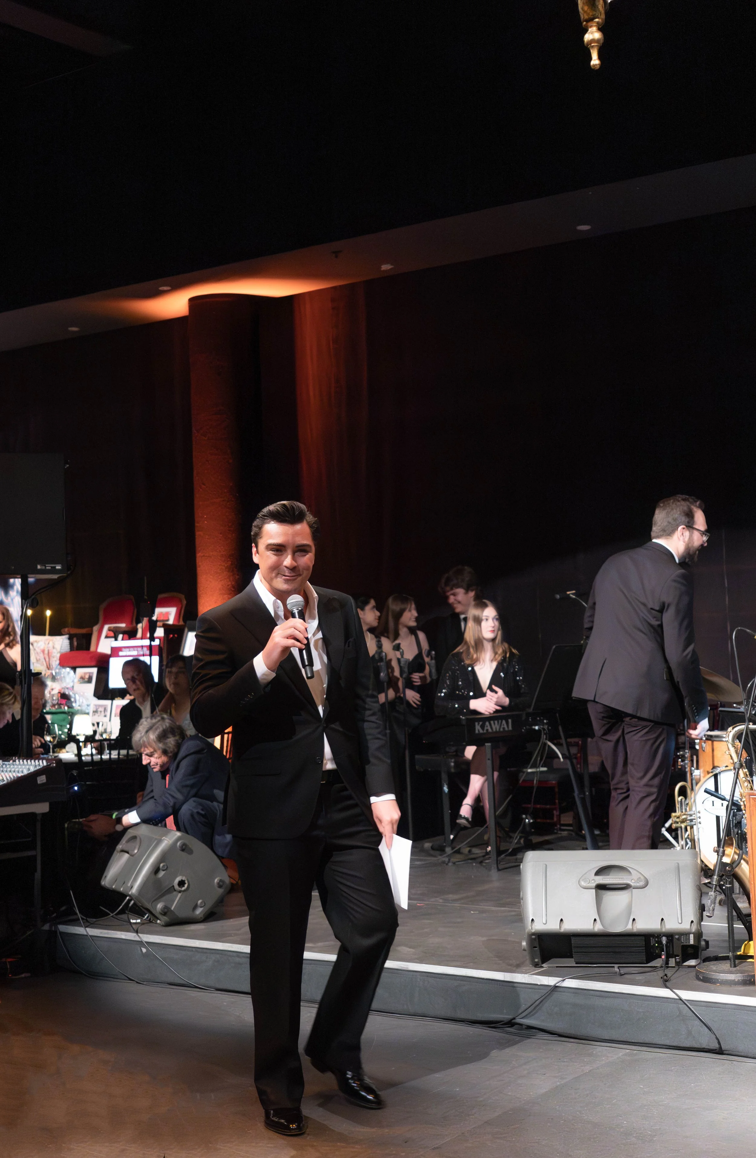 A male host wearing a black tuxedo and white shirt holding a microphone stands on stage at a formal event with a band playing in the background.