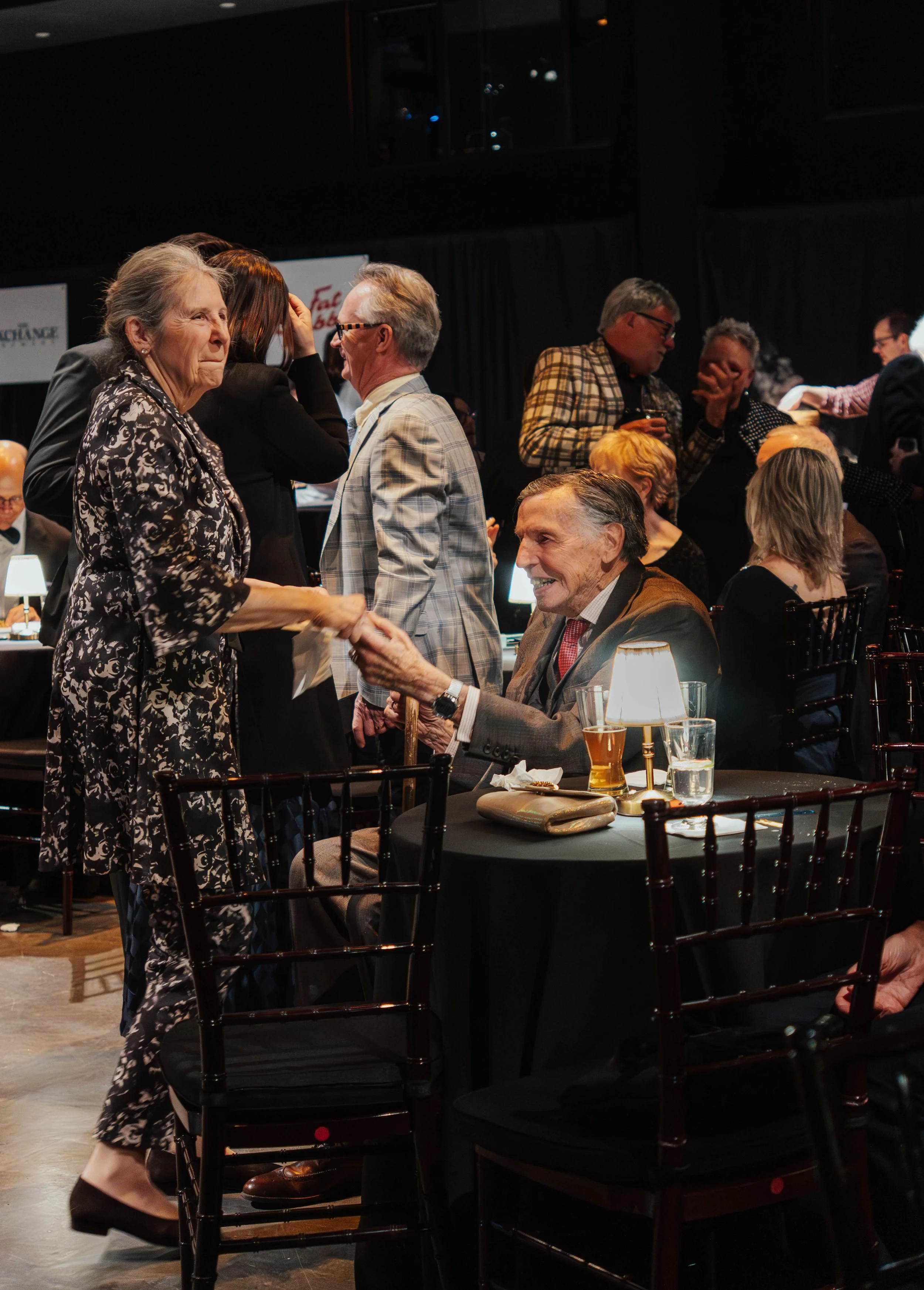Older man with gray hair smiling and shaking hands with a woman in a floral dress at a dinner event, surrounded by other people sitting at tables in a dimly lit room.