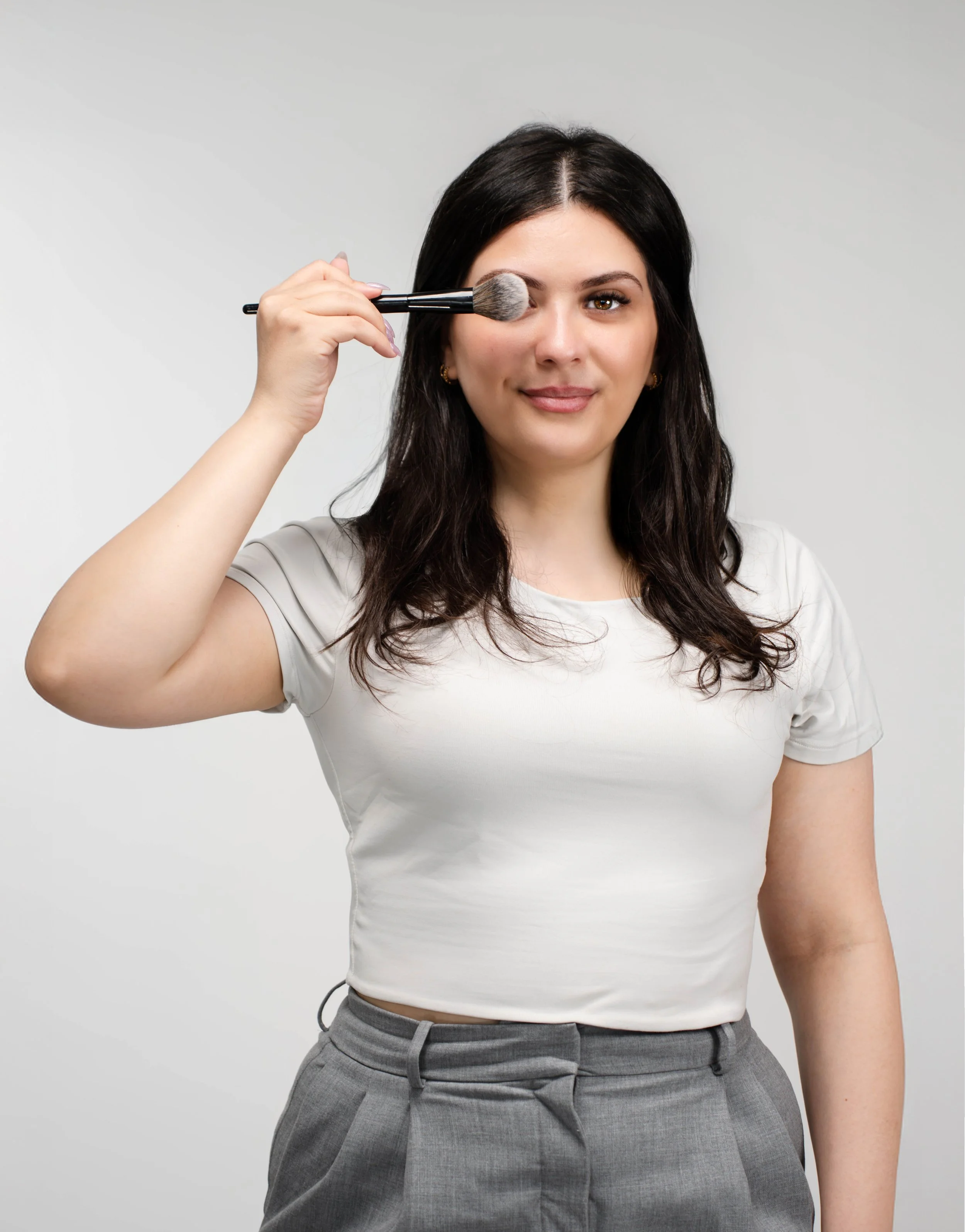 A woman with long dark hair wearing a white t-shirt and gray pants holding a makeup brush near her face against a plain gray background.