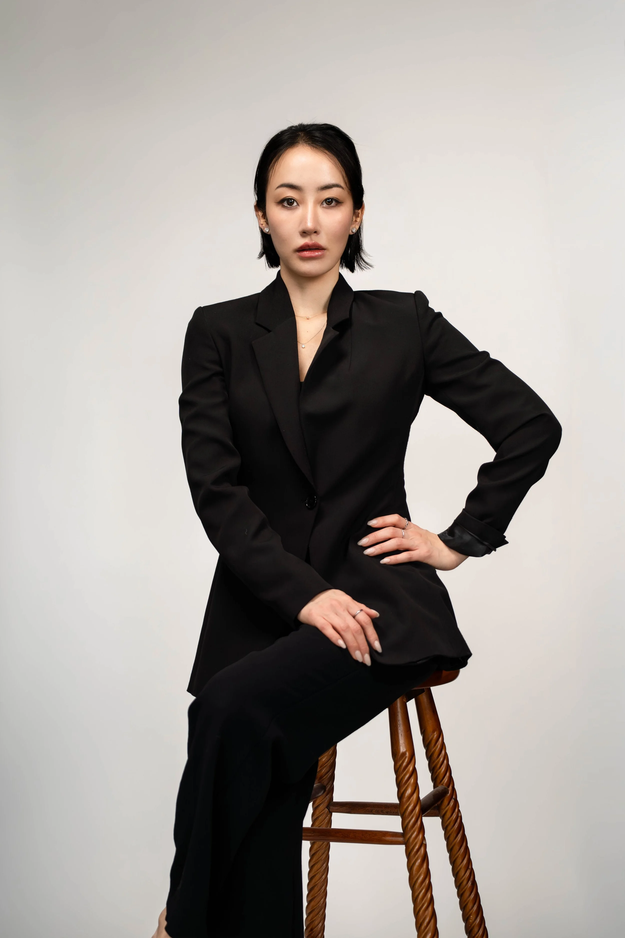 A woman with black hair and wearing a black suit, sitting on a wooden stool against a plain light background, looking directly at the camera.