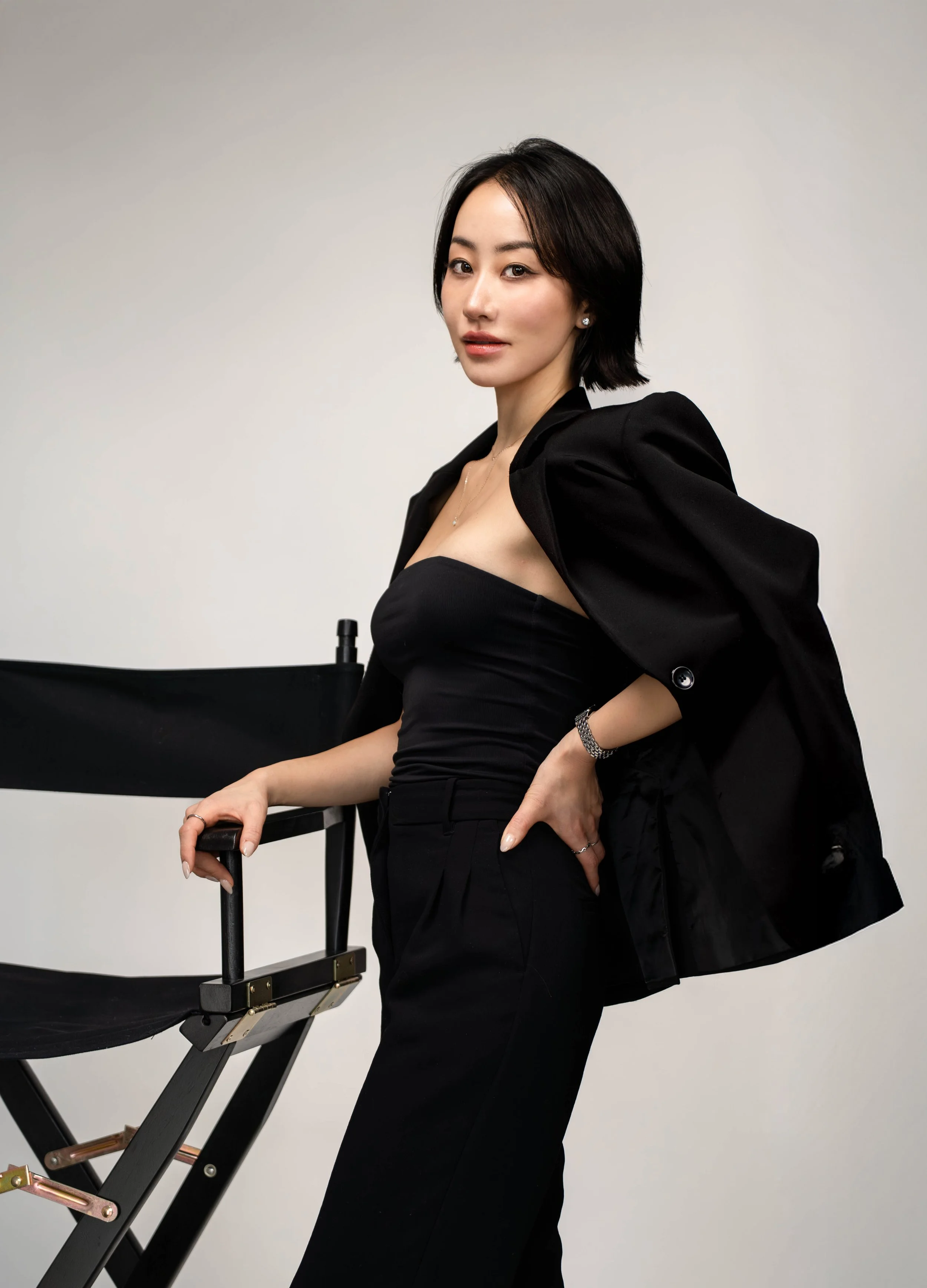 A woman in a black strapless top and black pants with a black jacket draped over her shoulders, standing next to a black director's chair, posing against a plain light-colored background.