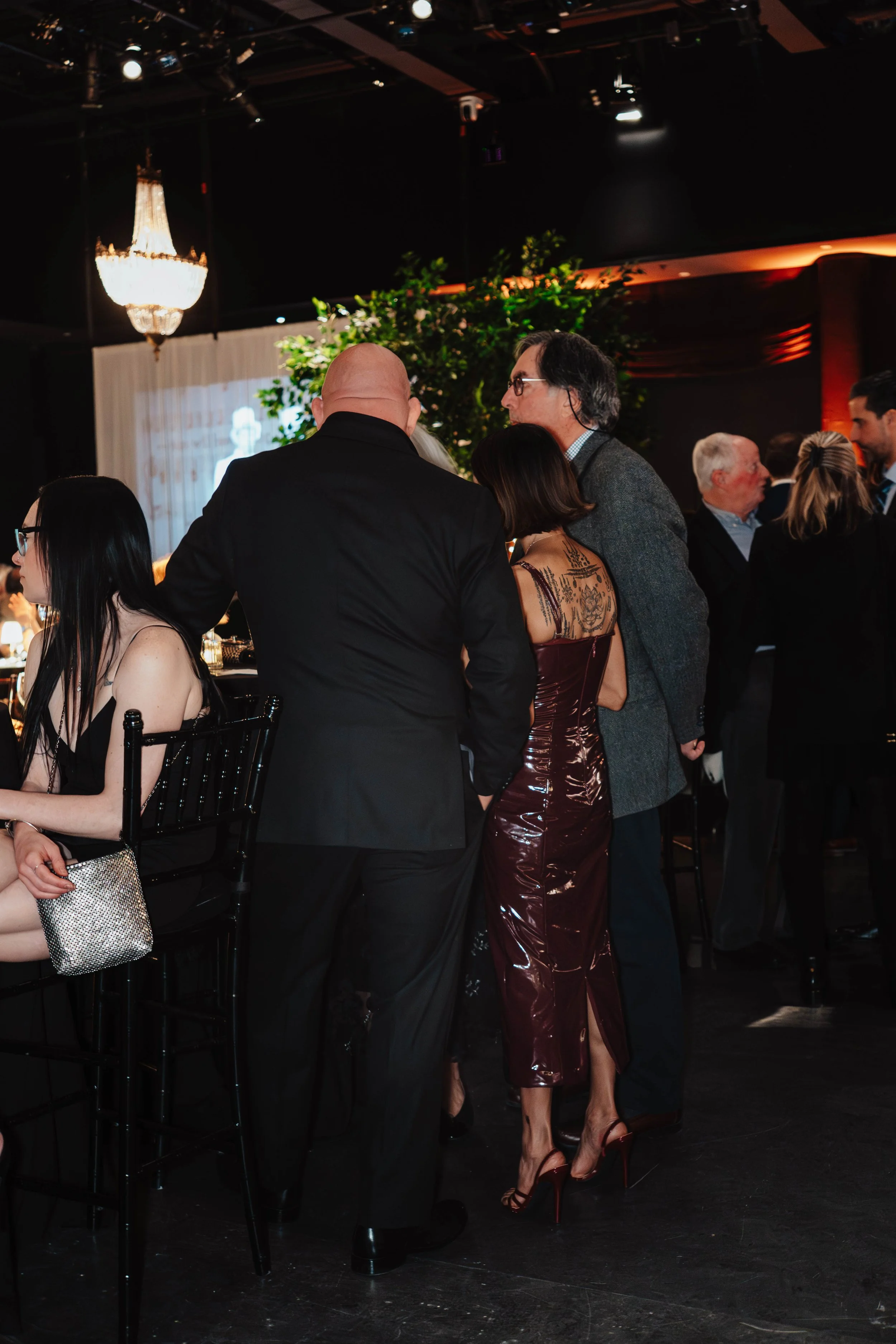 People socializing at an indoor event or party with a chandelier and decorative plants in the background.