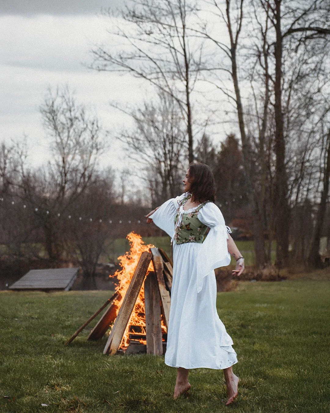 A woman in a white and green dress with white sleeves is barefoot on a grassy field, performing a dance or ritual near a small fire built from wooden planks outdoors during daytime.