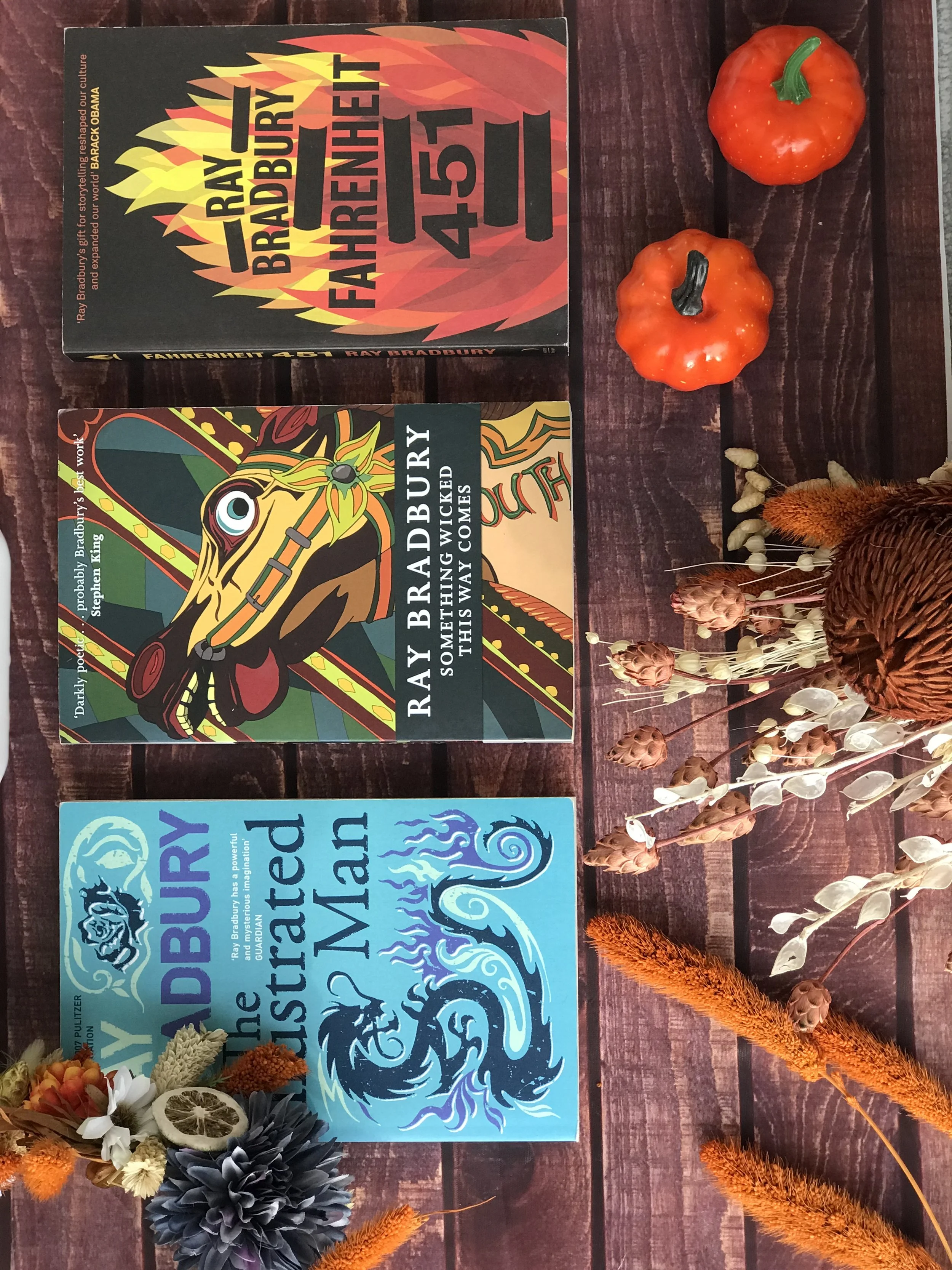 Three books by Ray Bradbury titled "Fahrenheit 451," "Something Wicked This Way Comes," and "The October Country" are laid out on a wooden surface. Two ceramic decorative pumpkins, one orange and one red, are positioned above the books. Dried flowers