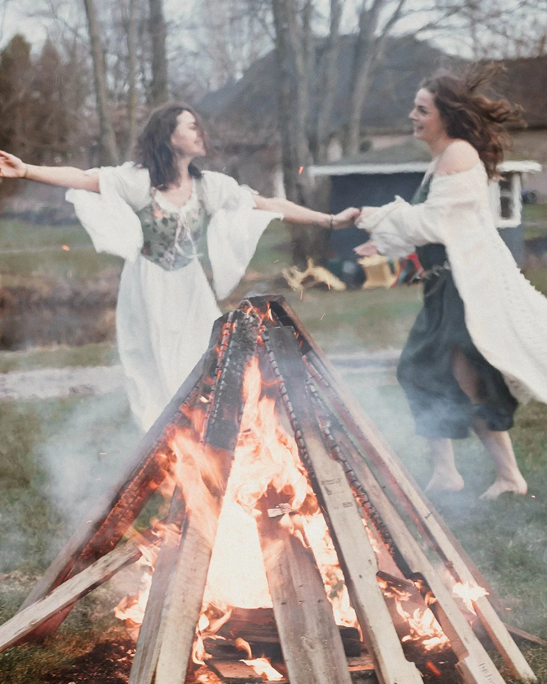 Two women dancing around a bonfire in an outdoor yard during dusk, holding hands and smiling, dressed in flowing white and dark skirts.