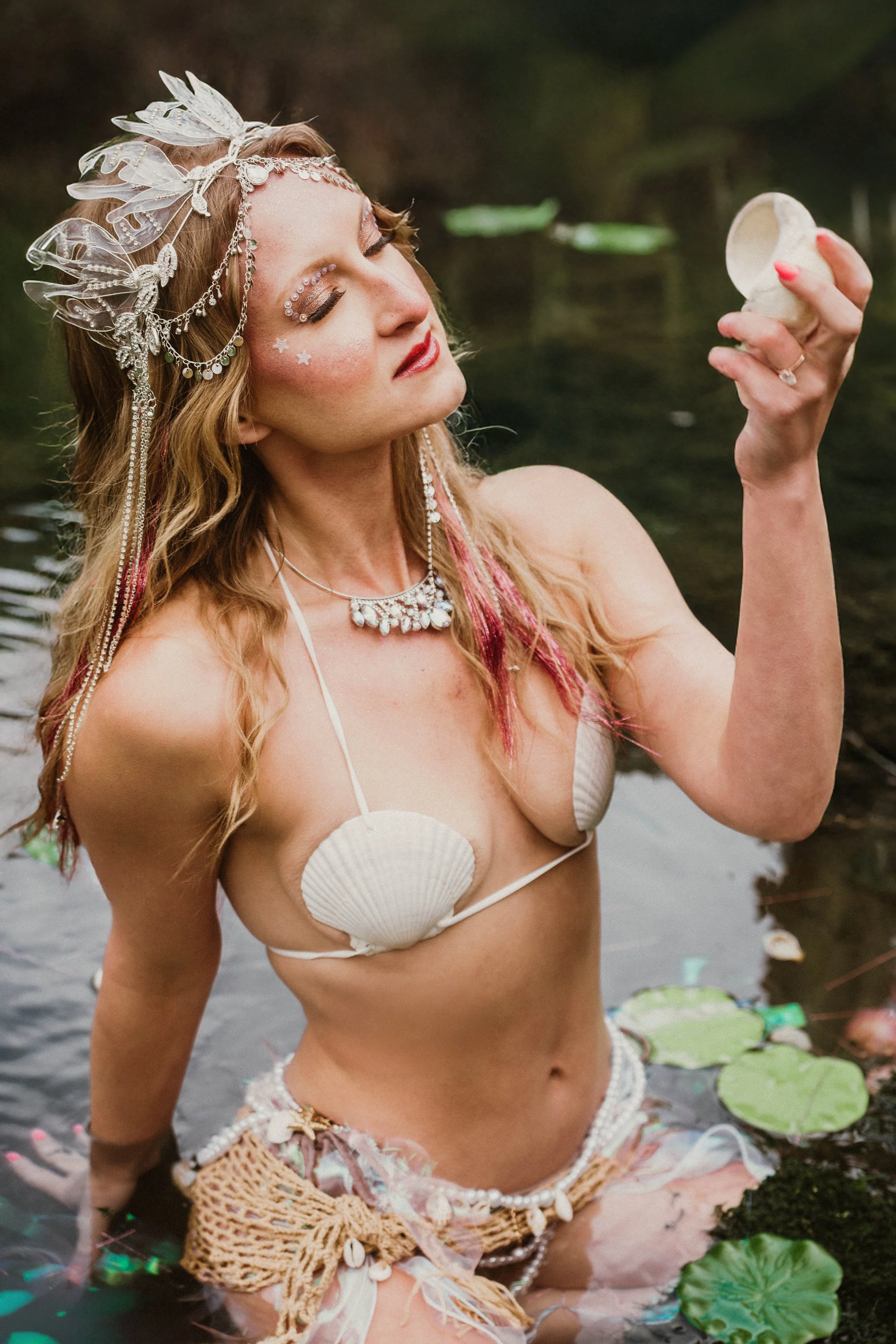 A woman with freckles and blonde hair, wearing a seashell top, jewelry, and a decorative headpiece with leaves, is standing in a pond with lily pads. She looks contemplative and is holding a small shell in her right hand.