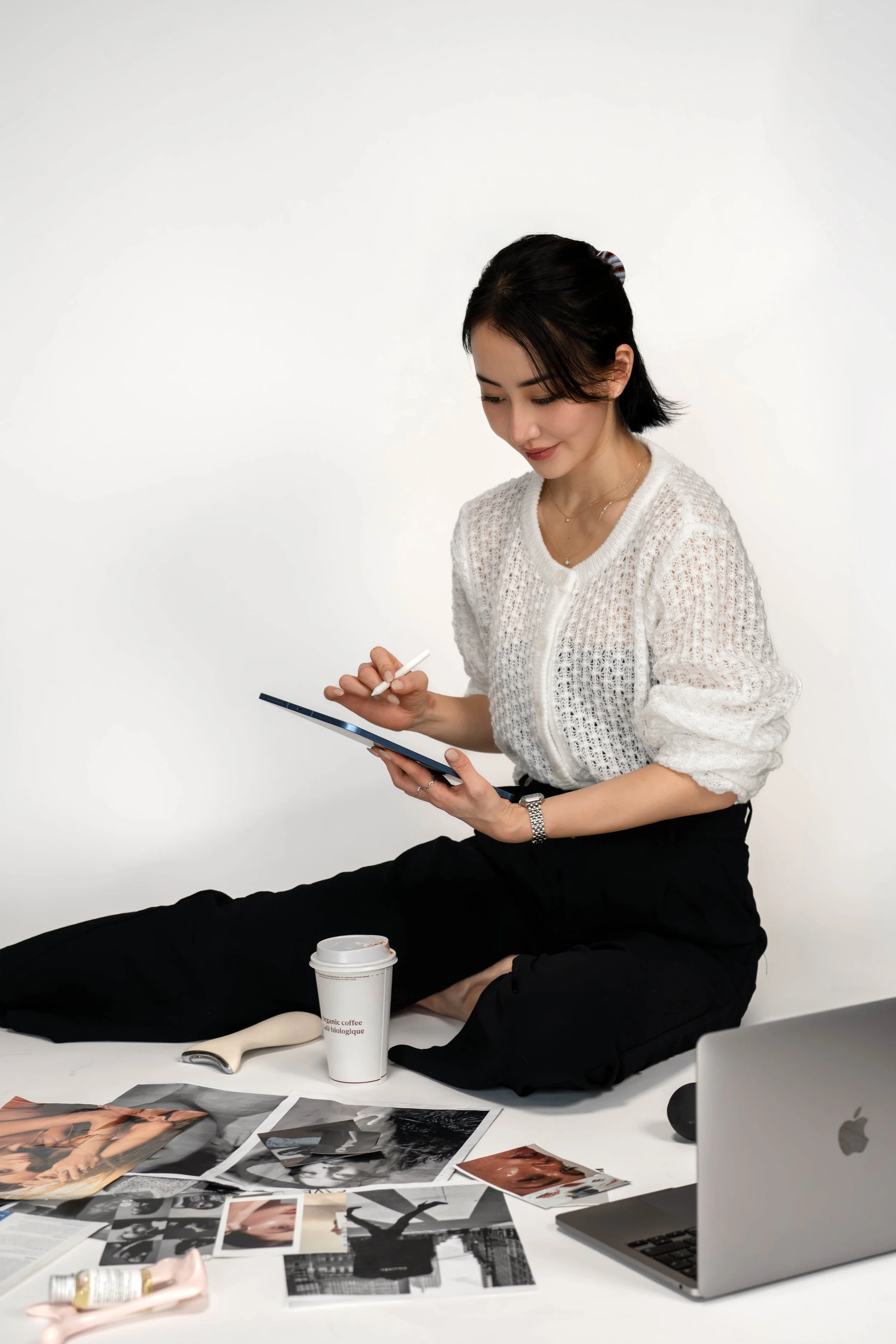 A woman sitting cross-legged on the floor, using a tablet with a stylus. She has short dark hair, is wearing a white crochet top and black pants, and has a silver watch. There are photos, a laptop, a coffee cup, and various items on the floor around 