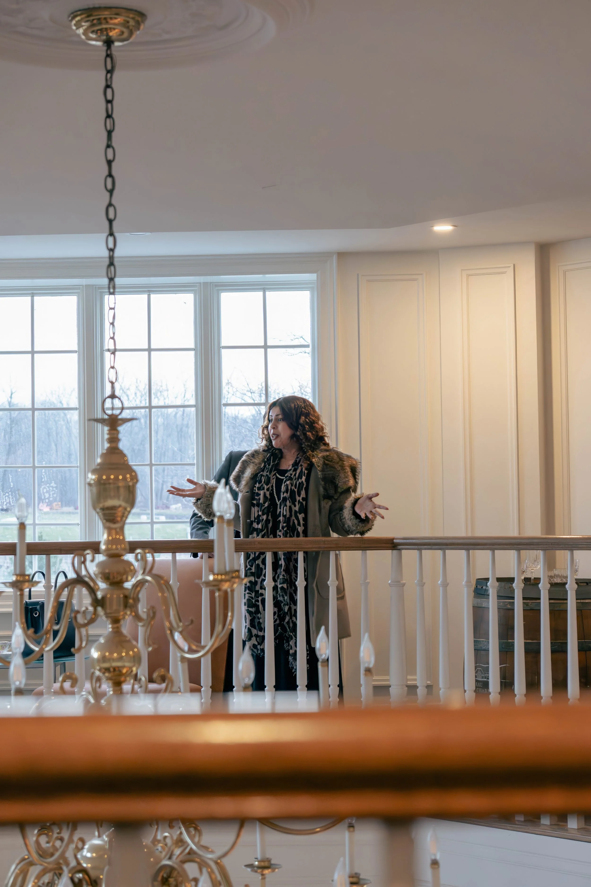 Woman with curly hair wearing a patterned scarf and coat, standing indoors near a white railing, speaking or gesturing, with large windows showing a scenic outdoor view in the background.