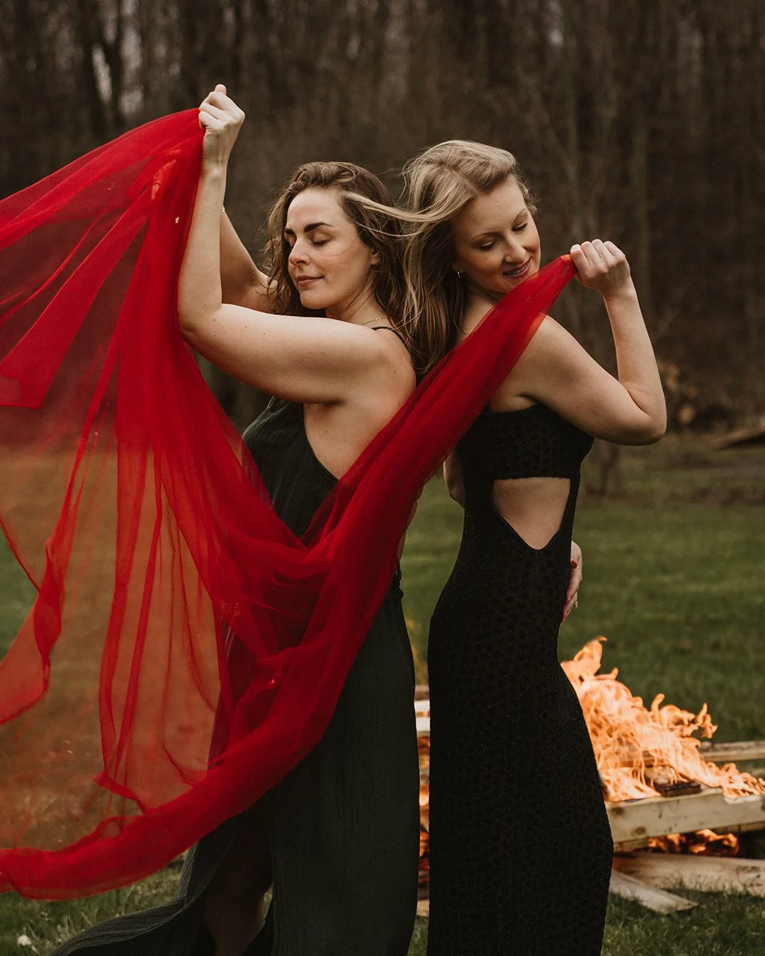 Two women dressed in black dresses, standing back to back outdoors, holding red flowing fabric, with a fire and a pile of wood in the background.
