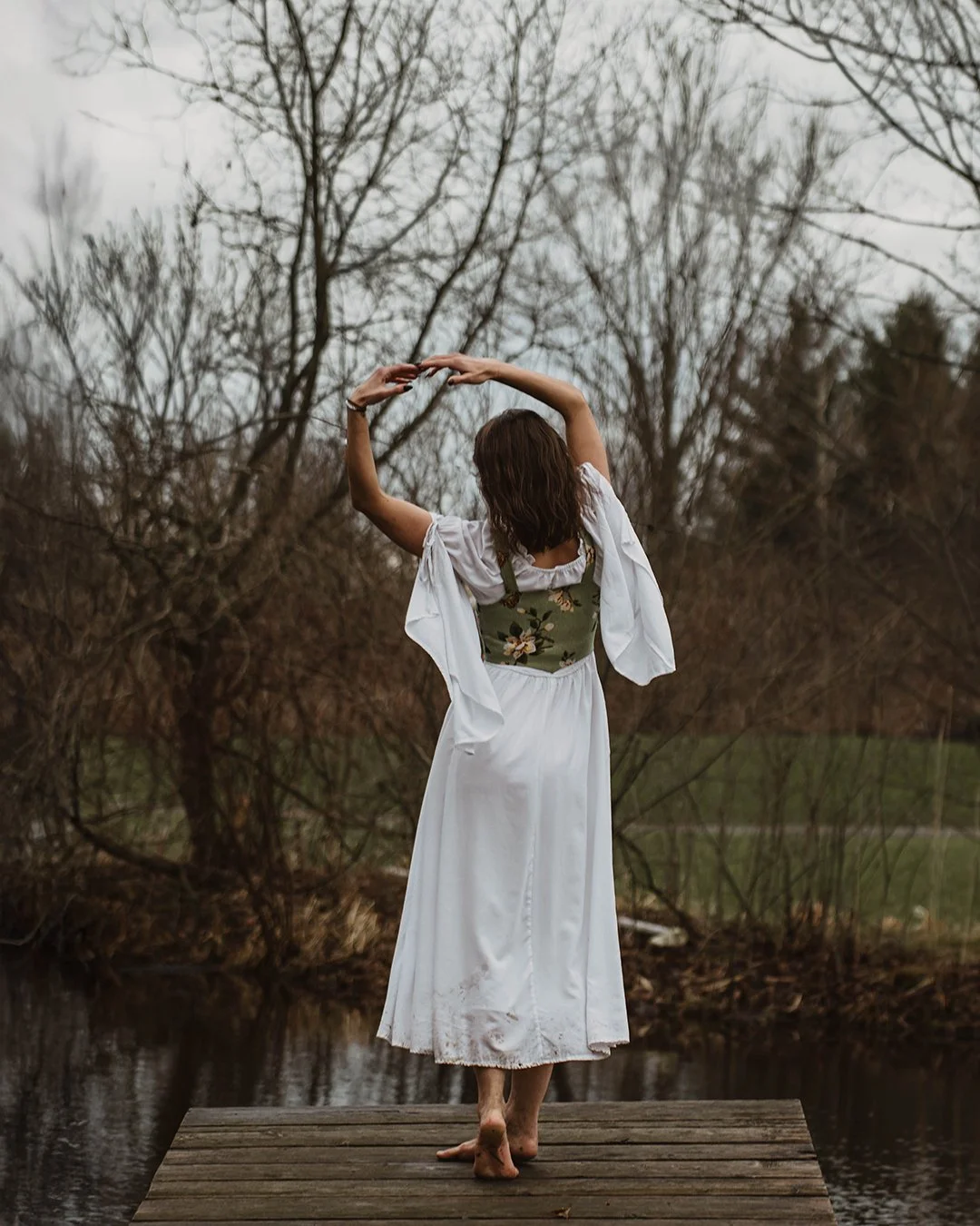 Woman standing on a wooden dock near water, with her back to the camera, arms raised above her head, surrounded by leafless trees during overcast weather.