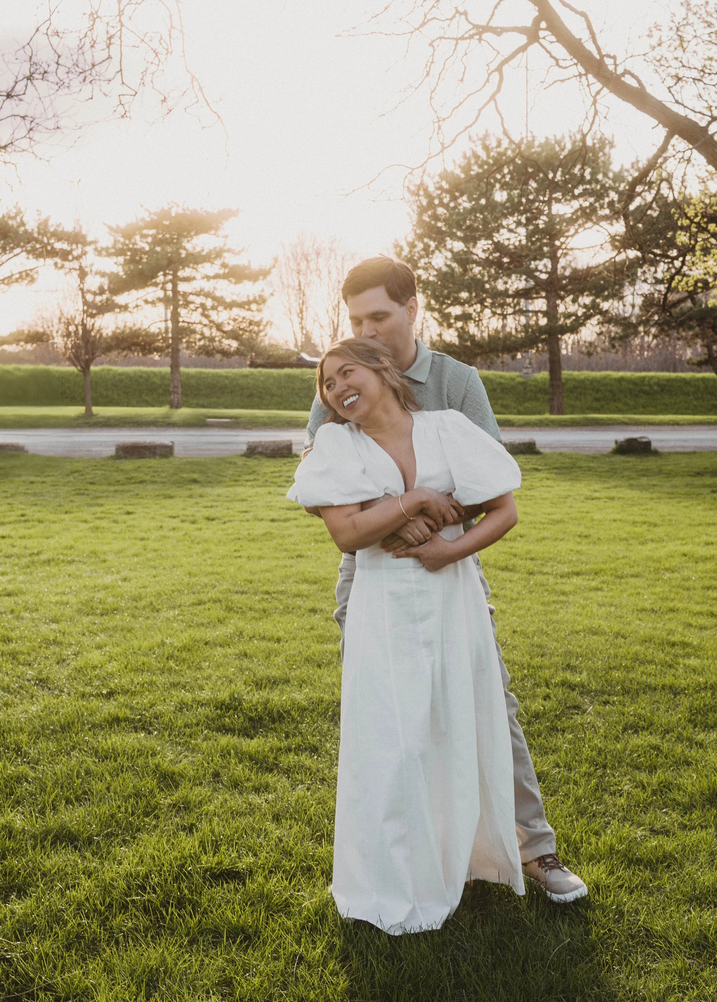 A smiling woman in a white dress hugging a man from behind, both standing on green grass in a park with trees and a pathway, during sunset.