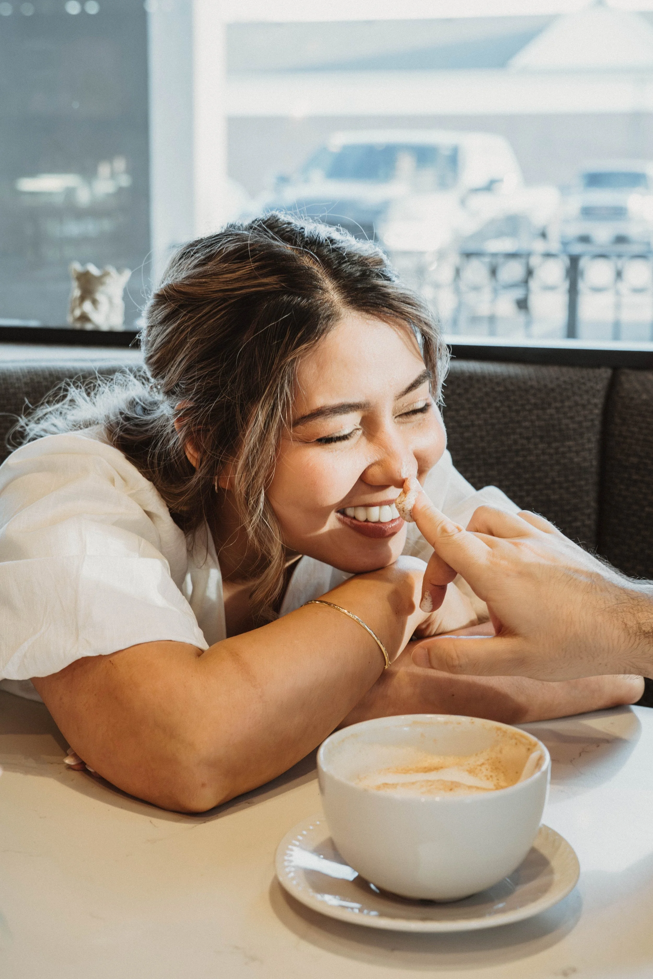 A woman smiling with her eyes closed, resting her head on her folded arms on a table, with a cup of coffee in front of her. She is wearing a white shirt and a gold bracelet. In the background, there is a large window with a parking lot visible outsid