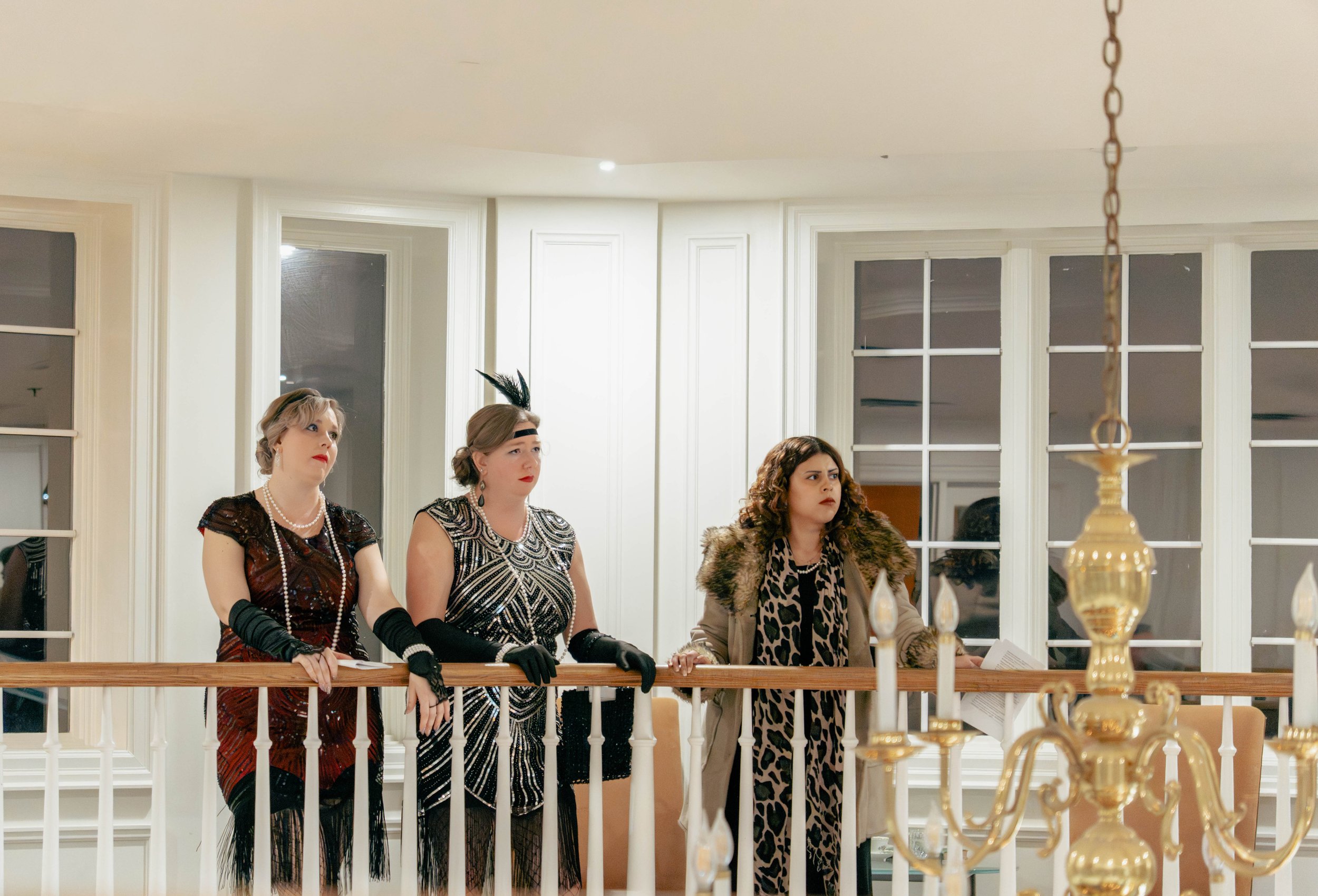 Three women dressed in vintage 1920s style clothing standing on a balcony with white panel walls and large windows, looking in the same direction with serious expressions.
