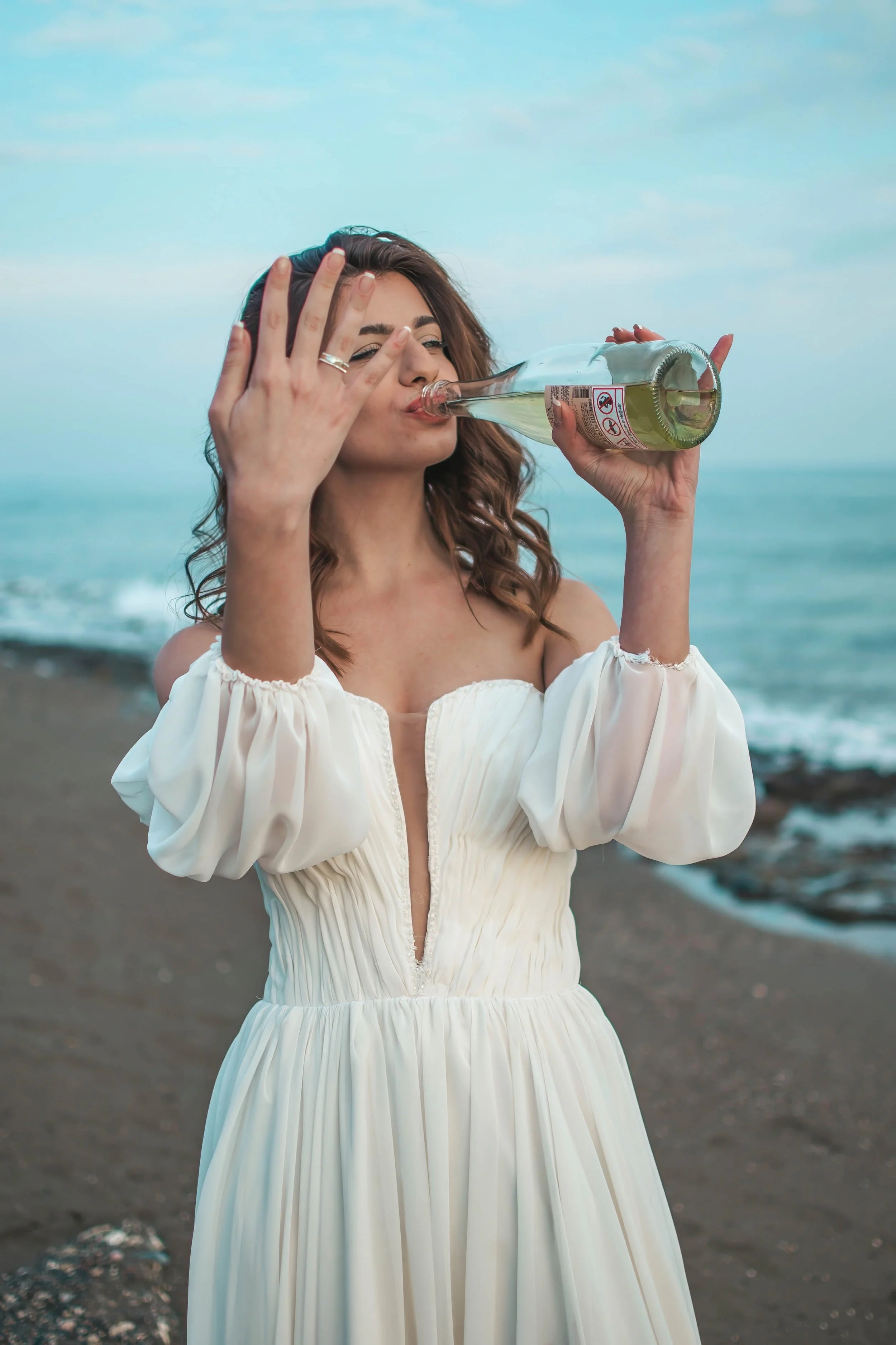 Woman in white dress drinking from a glass bottle on a beach with ocean in the background.