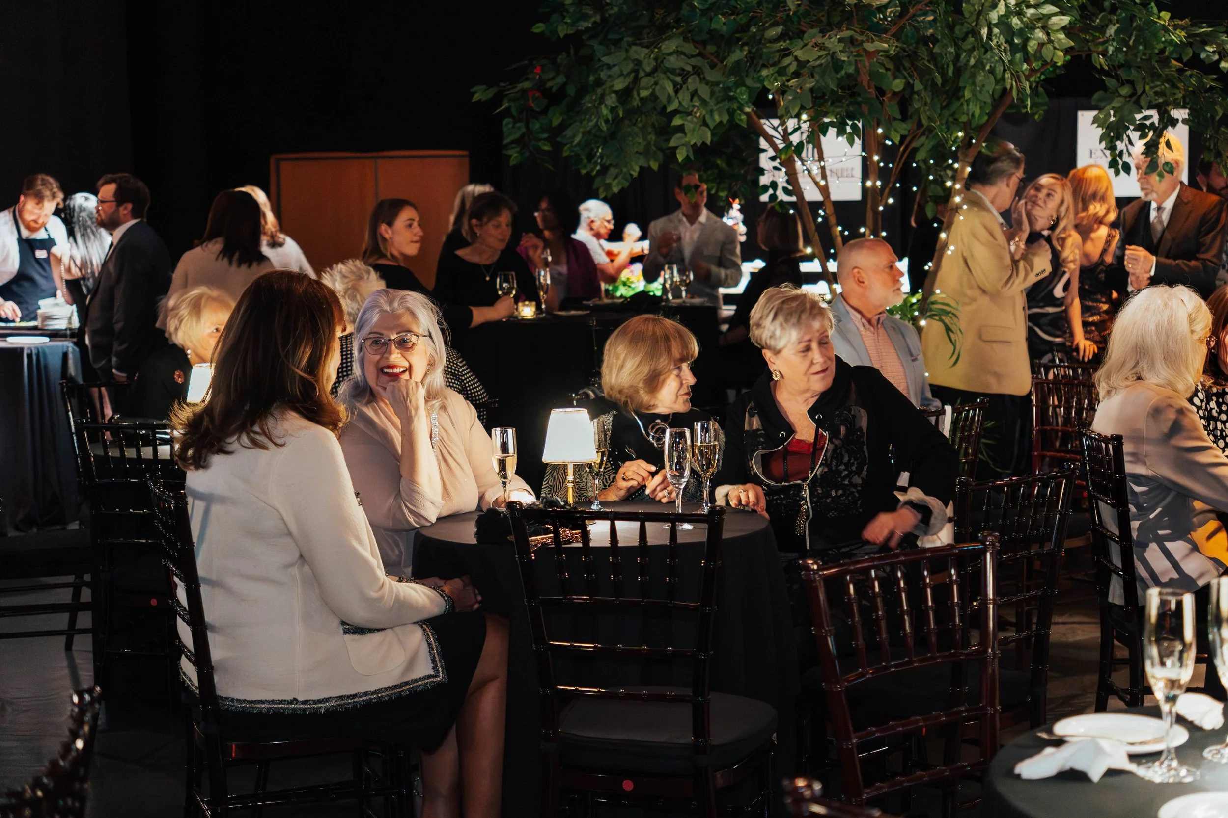 People gathered at a formal event or banquet, sitting at round tables with black tablecloths, glasses of champagne, and small lamps, under a decorated tree with string lights.