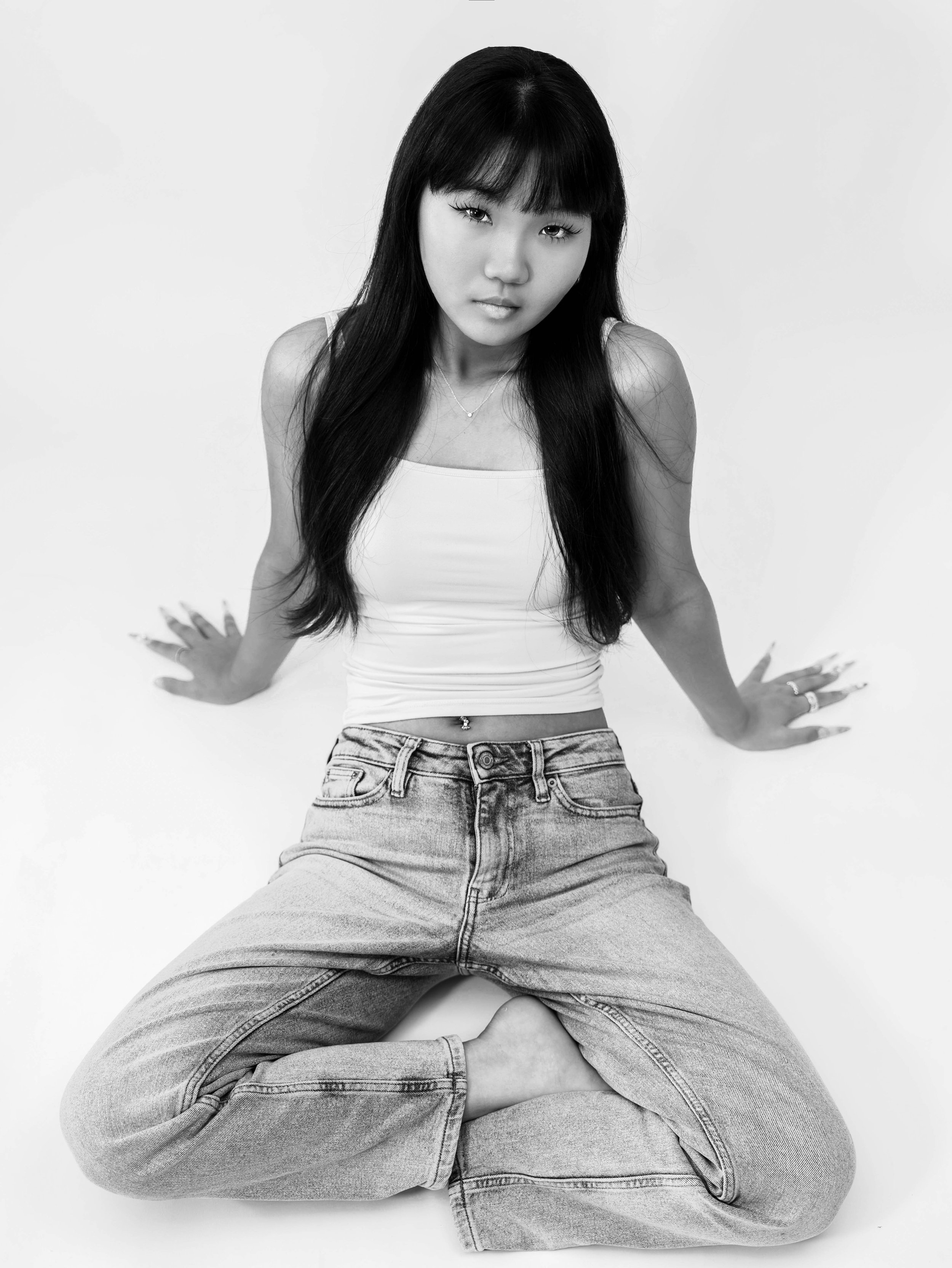 Black and white editorial studio portrait of Dana seated against a white backdrop, wearing a white tank top and jeans while looking directly at the camera, photographed by Lara Grace Creative Studio in Toronto.