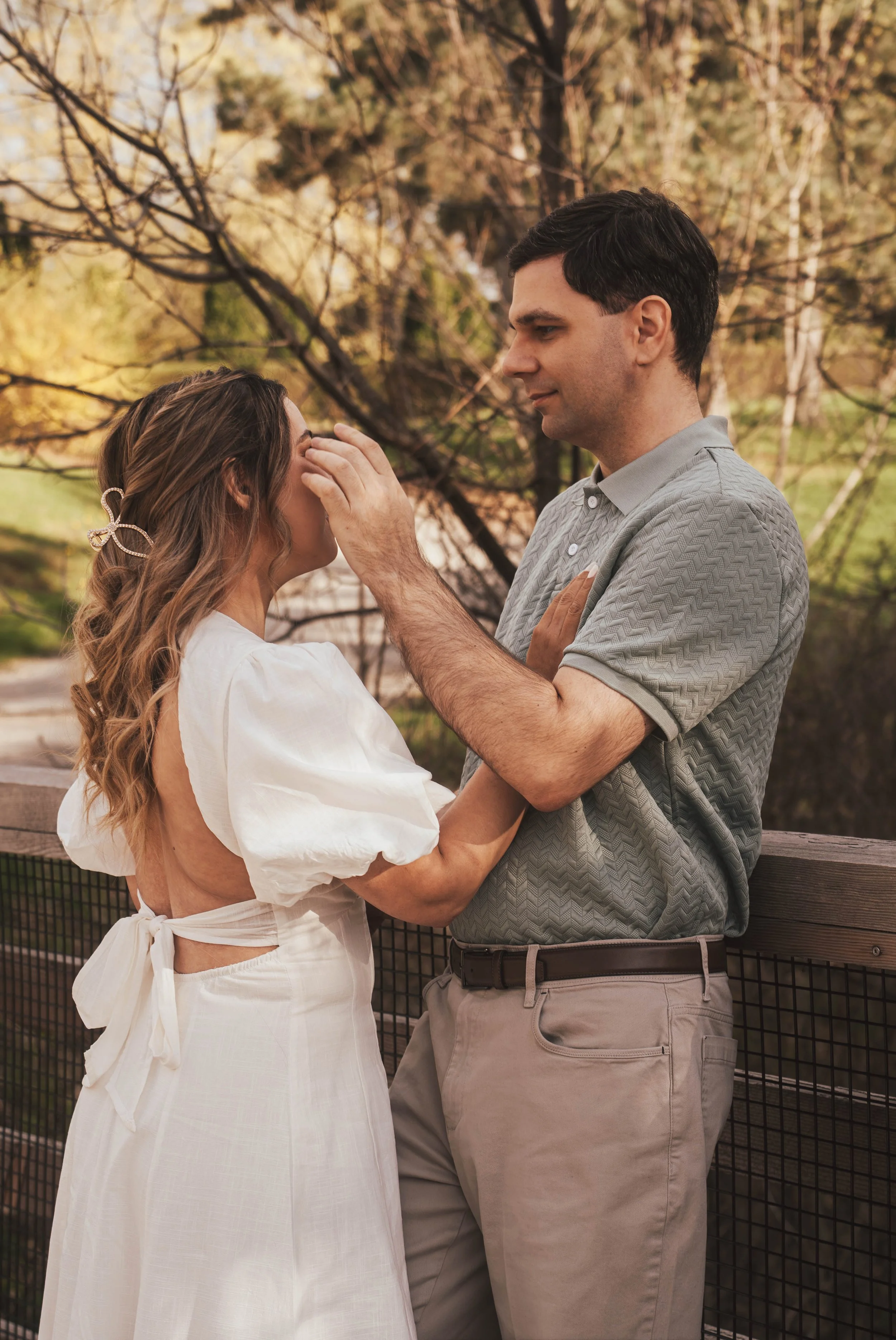 A man and a woman standing close together outdoors during autumn, with trees with yellow and brown leaves in the background. The man is gently touching the woman's forehead, and their eyes are closed, sharing an intimate moment.