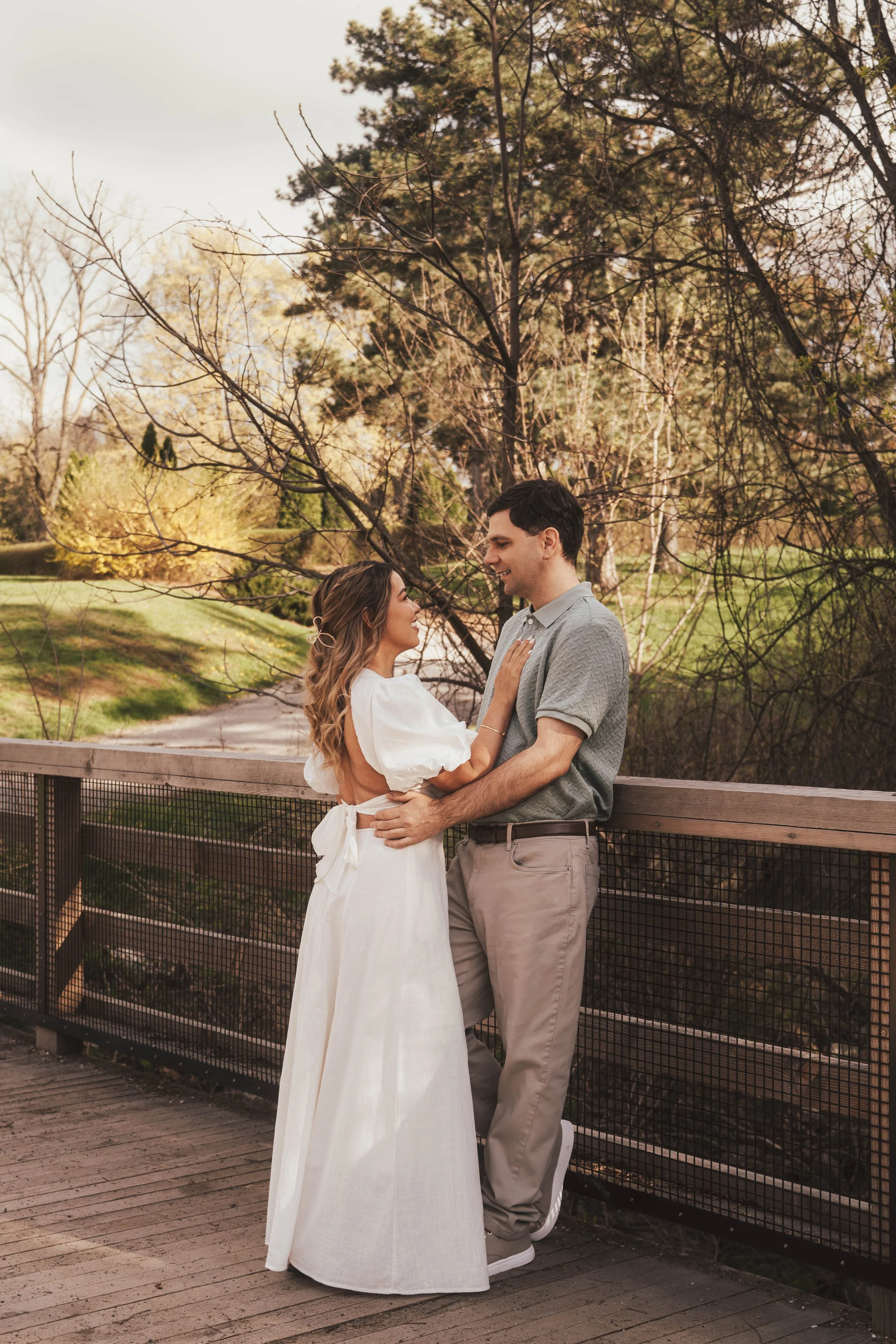 A couple standing closely together outdoors, with the woman touching the man's chest and both smiling at each other. They stand on a wooden bridge with a black railing, surrounded by trees with sparse foliage in the background. The woman wears a whit
