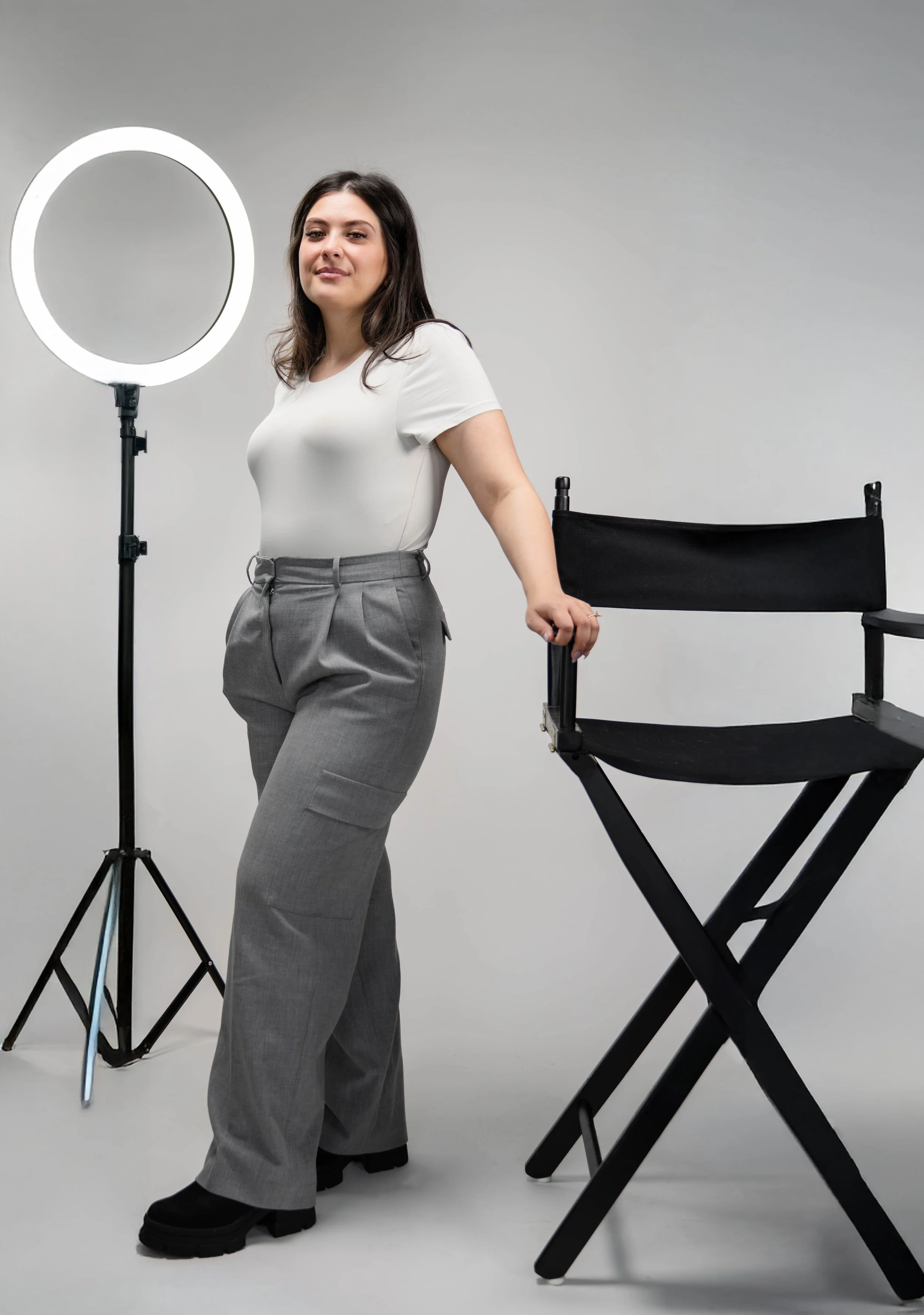 A woman with dark hair, wearing a white t-shirt and gray pants, stands in a photography studio with a ring light and a black director's chair.